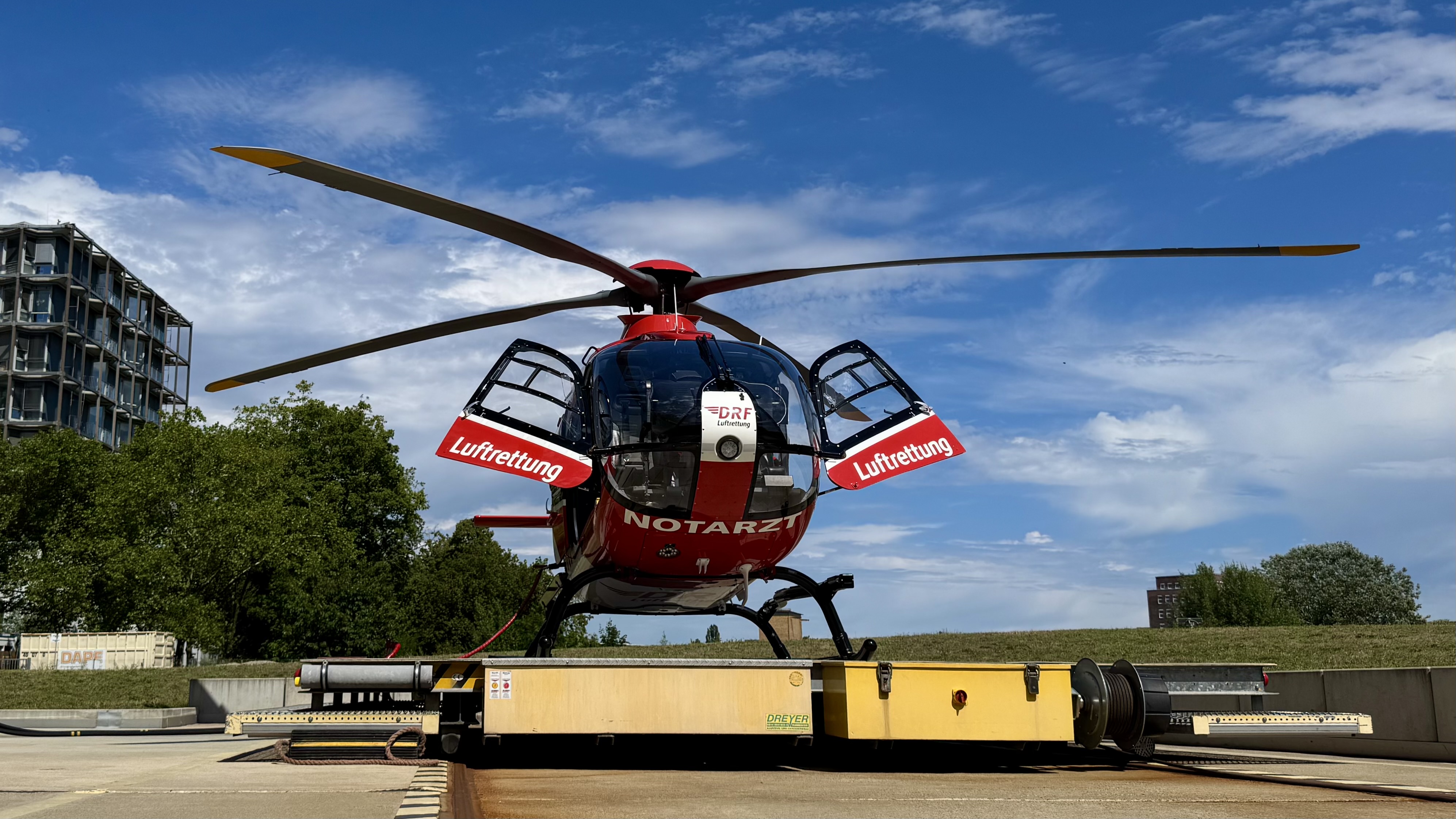 Ein Rettungshubschrauber steht mit geöffneten Türen auf einem Landeplatz. Er ist rot mit Aufschriften für "Luftrettung" und "Notarzt". Im Hintergrund sind Bäume und Gebäude sichtbar unter einem klaren blauen Himmel.