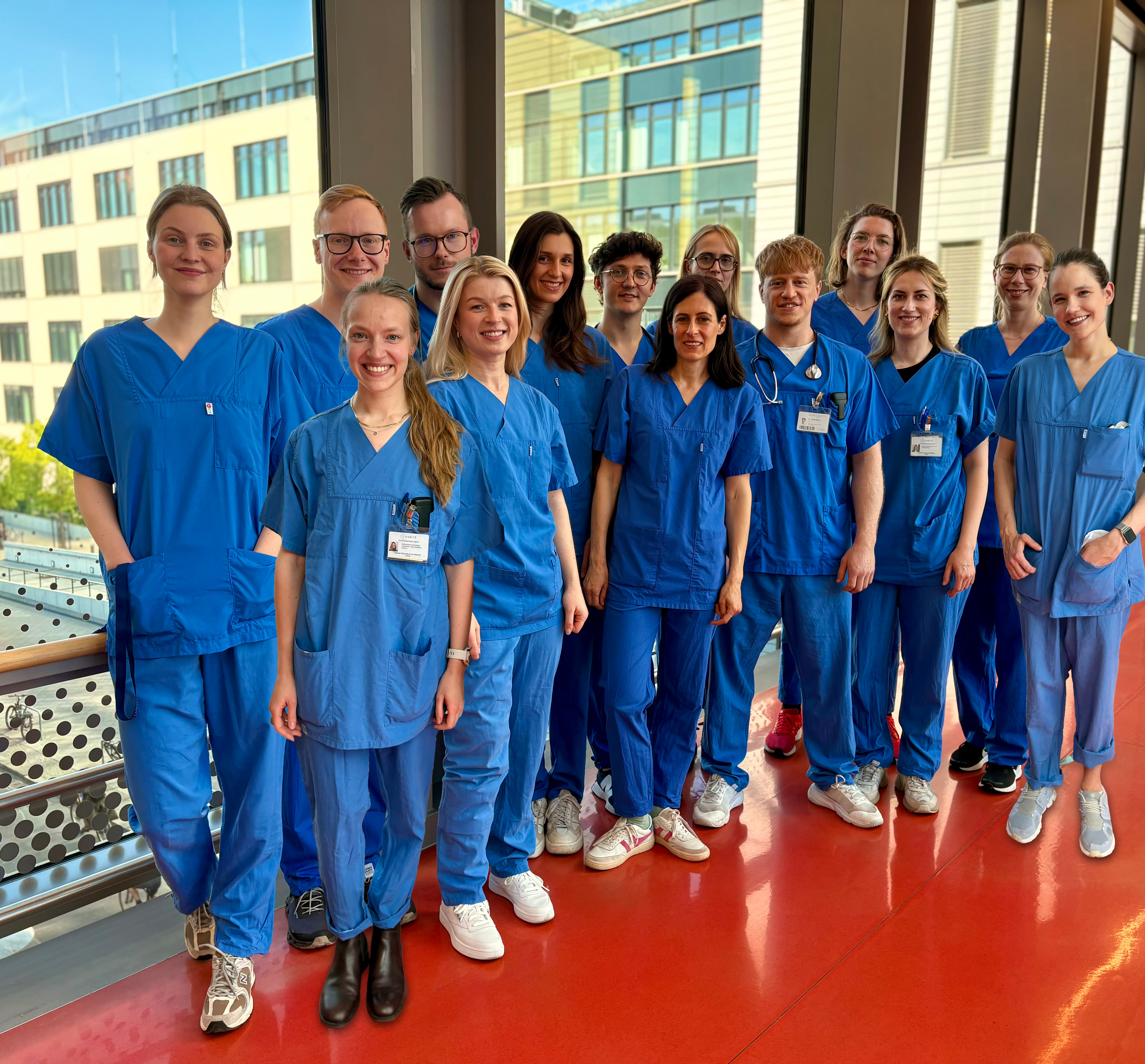 Eine Gruppe von 15 Personen in blauen Klinikoberteilen und -hosen steht lächelnd in einem Raum mit großen Fenstern. Im Hintergrund sind moderne Gebäude sichtbar. Die Szene vermittelt eine freundliche, professionelle Atmosphäre.