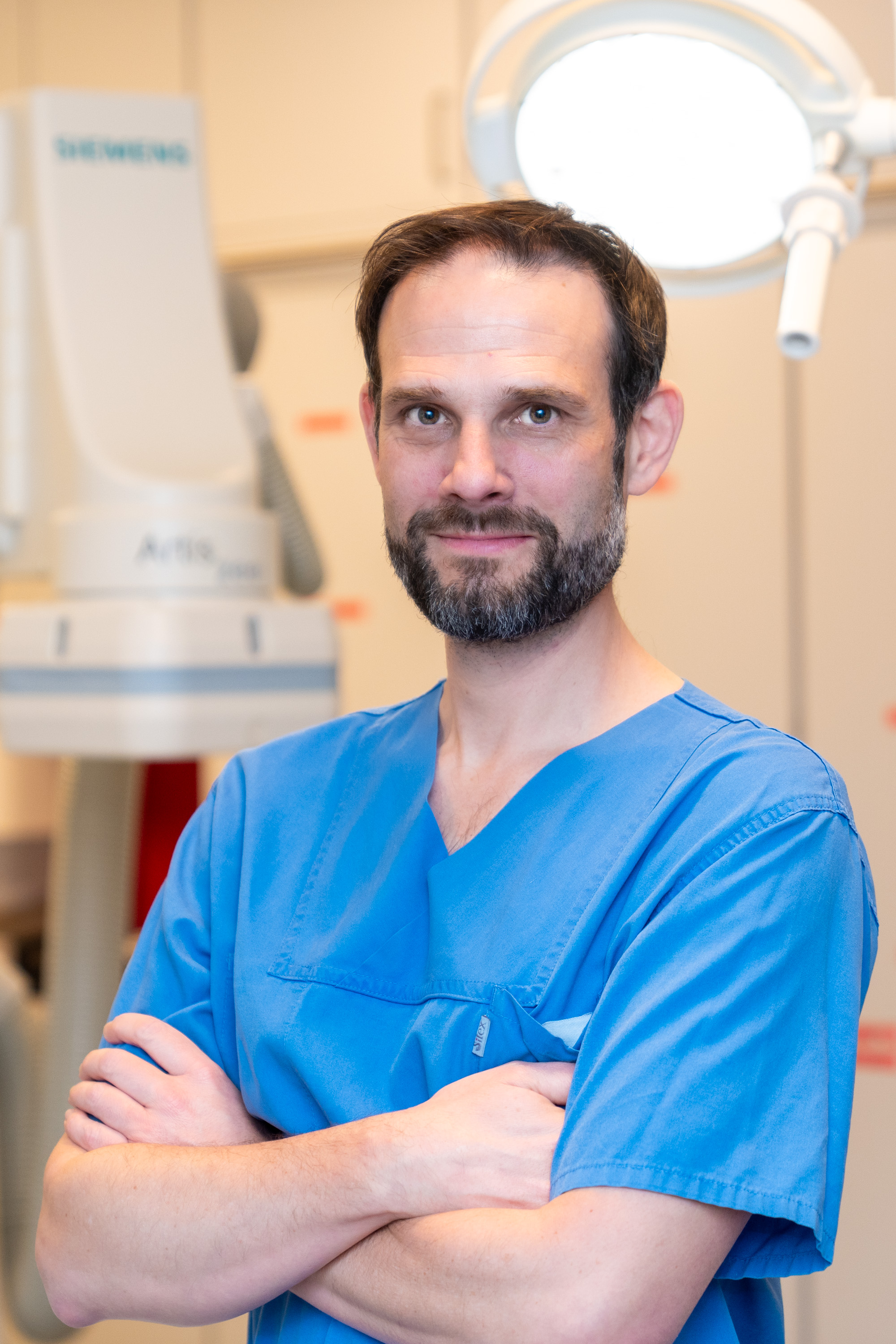 Ein Mann mit Bart und kurzer Frisur trägt ein blaues medizinisches Outfit. Er steht in einem Untersuchungsraum mit verschränkten Armen und einem freundlichen Ausdruck. Im Hintergrund ist ein medizinisches Gerät und eine Lampe sichtbar.