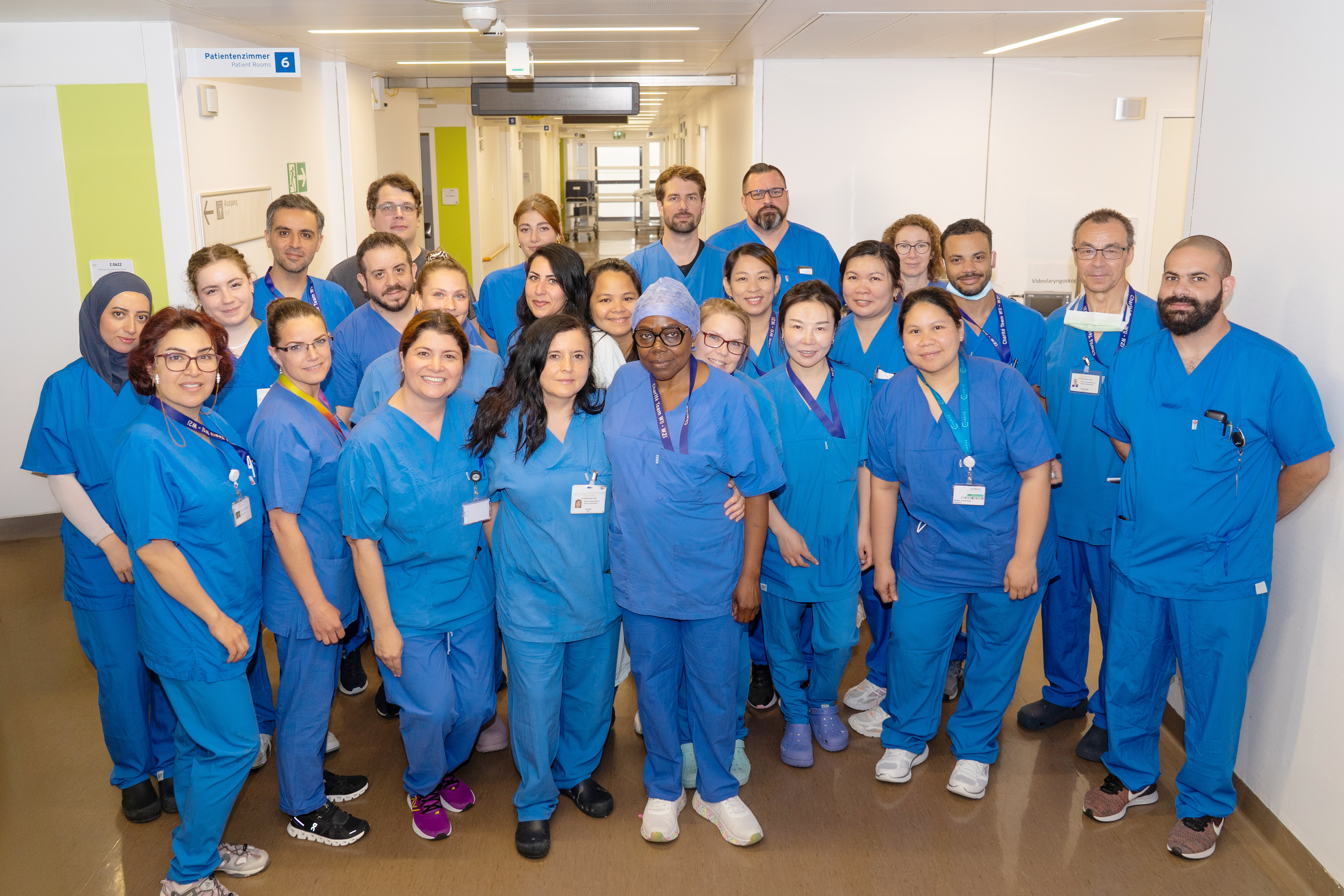 Eine Gruppe von 20 Personen in blauen Krankenhausuniformen steht in einem Flur. Alle lächeln und blicken in die Kamera. Im Hintergrund sieht man Symptome eines modernen Krankenhauses, wie Patientenzimmerschild und helle Wände.