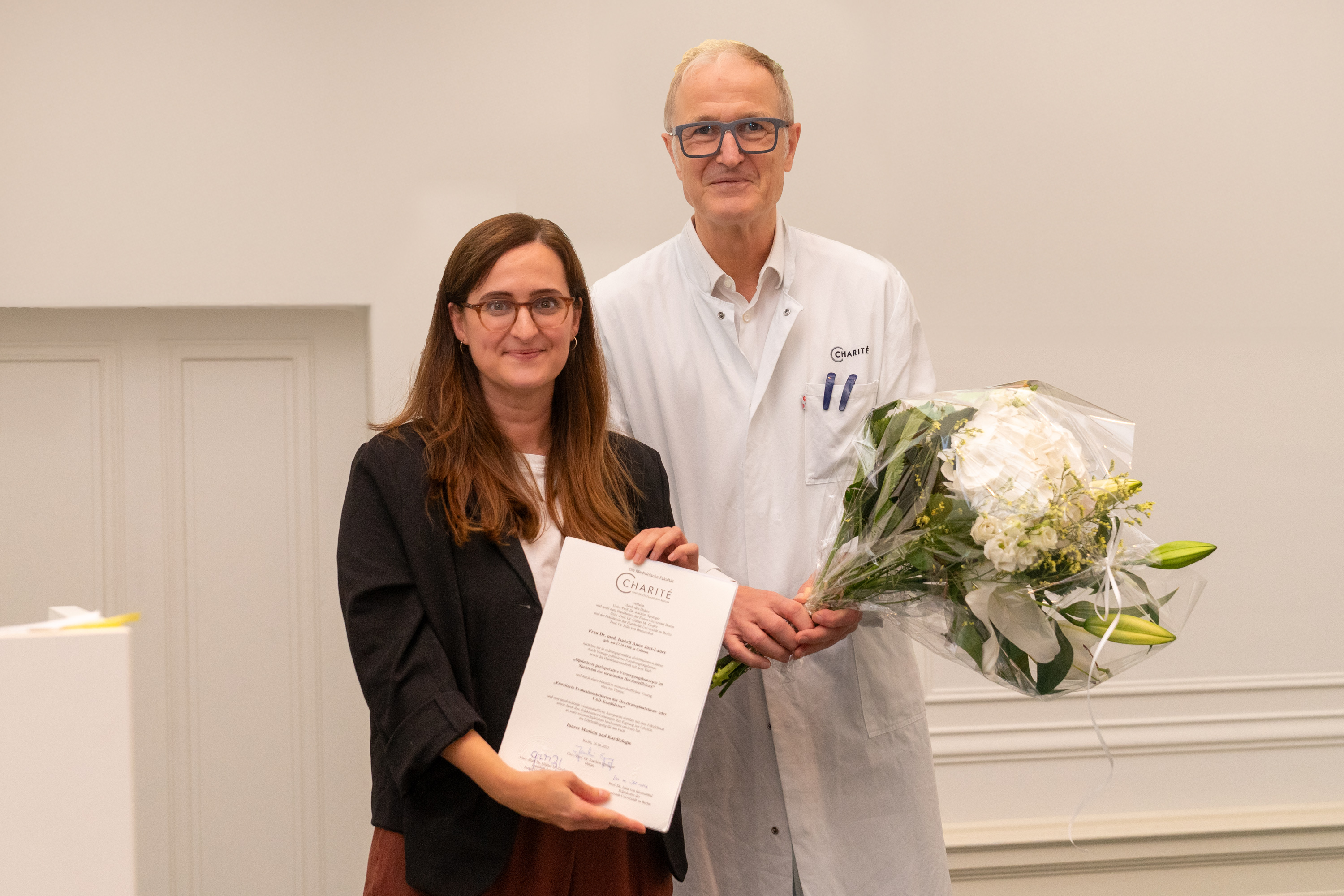 Eine Frau in einem Blazer hält ein Dokument und lächelt. Neben ihr steht ein Mann in einem weißen Arztkittel, der einen Blumenstrauß in der Hand hält. Beide stehen in einem hellen Raum, was auf einen feierlichen Anlass hindeutet.