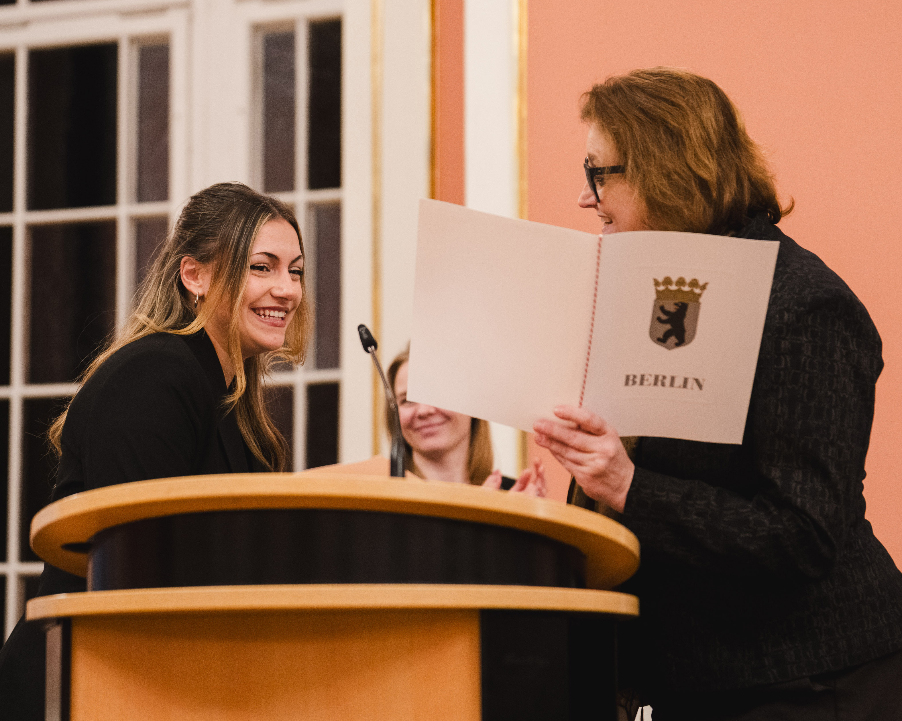 Eine lächelnde Frau erhält von einer anderen Frau eine Urkunde mit dem Stadtwappen von Berlin. Im Hintergrund stehen weitere Personen. Die Szene vermittelt eine feierliche Stimmung, möglicherweise eine Zeremonie oder Auszeichnung.