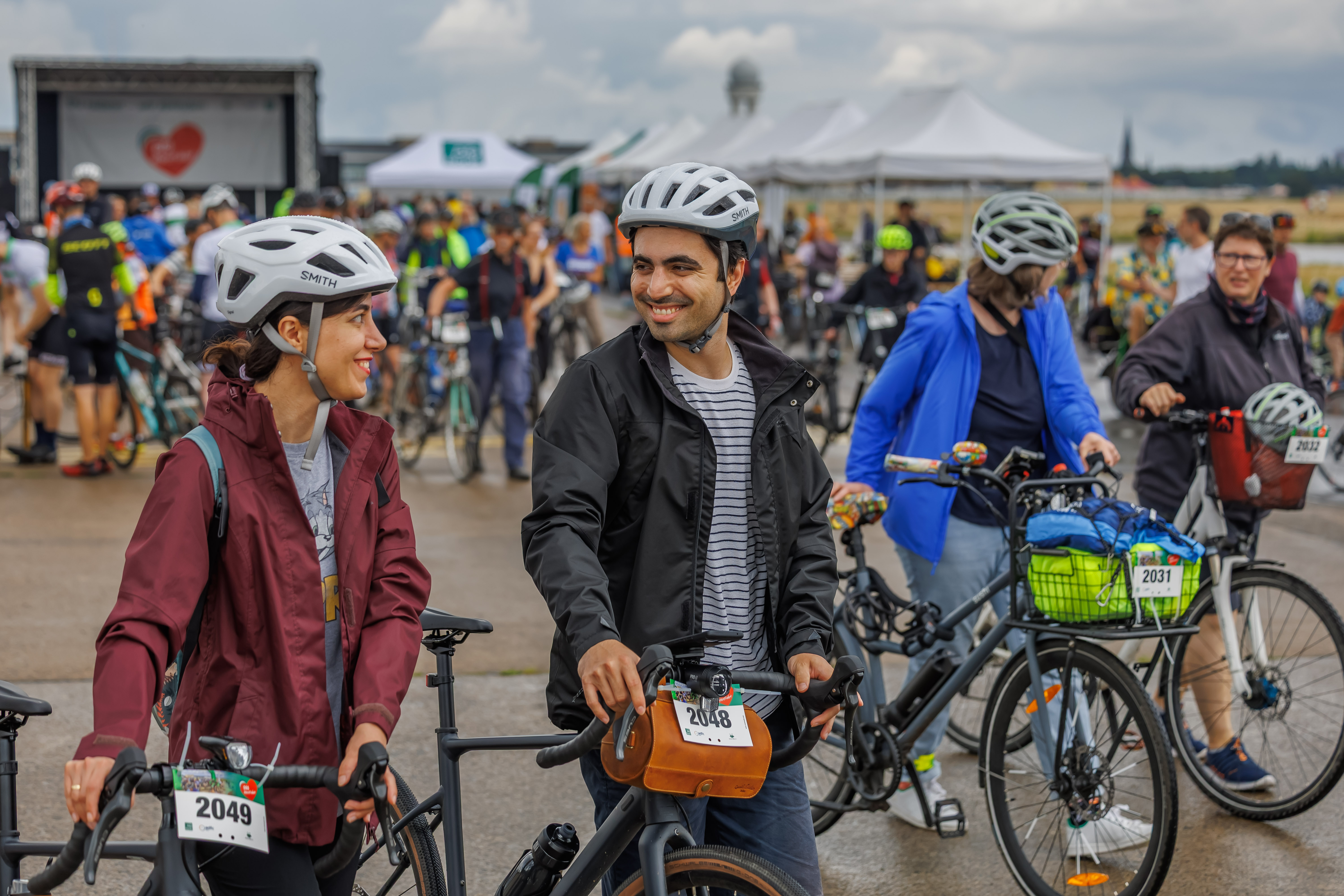 Zwei Radfahrer, eine Frau in einer roten Jacke und ein Mann in schwarzer Jacke, stehen lächelnd nebeneinander. Im Hintergrund sind weitere Teilnehmer mit Fahrrädern und Zelten zu sehen, während die Veranstaltung in einer belebten Umgebung stattfindet.
