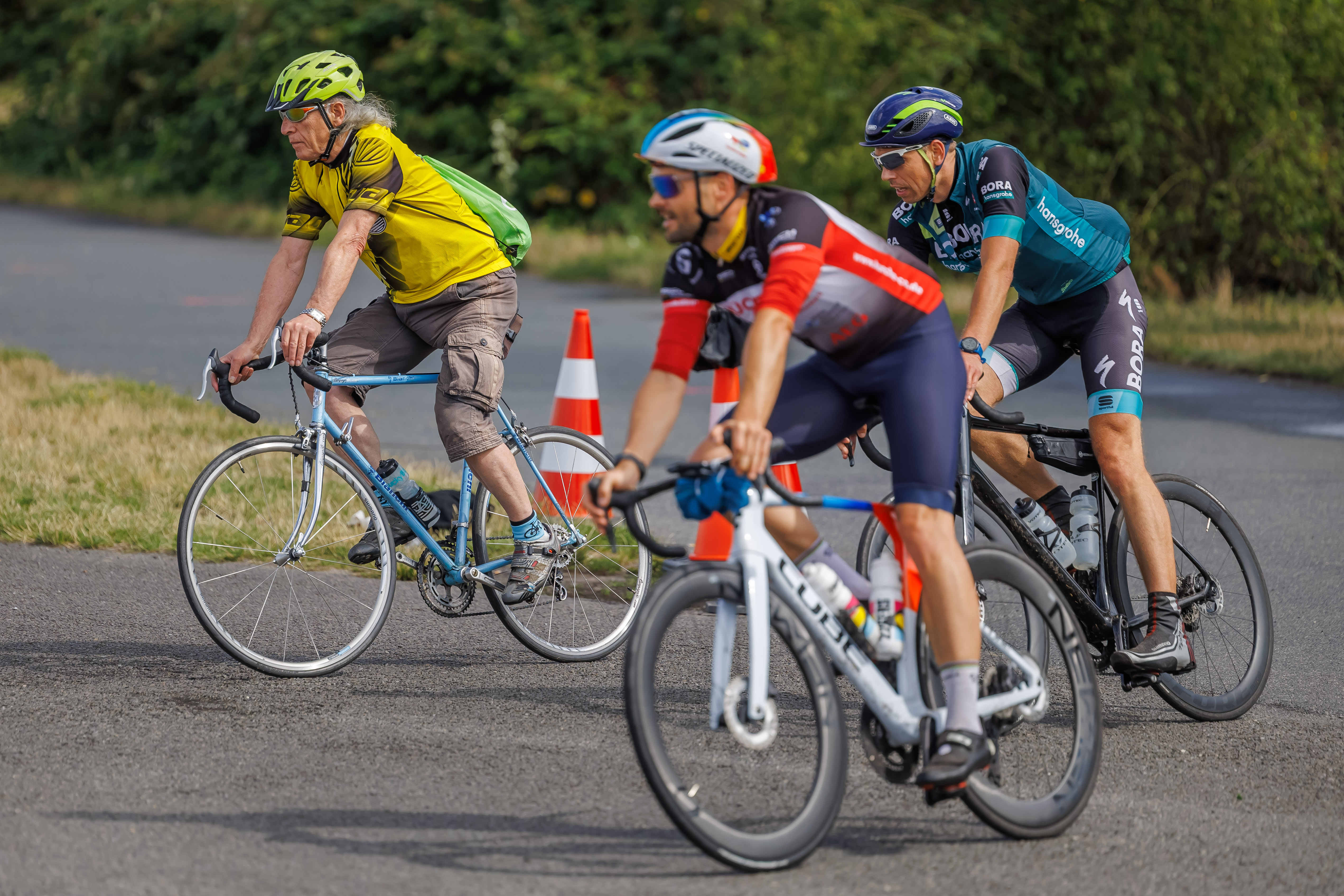 Drei Radfahrer fahren auf einer Straße. Ein älterer Mann in gelb trägt einen grünen Helm, während zwei jüngere Männer in sportlicher Radbekleidung mit angepasster Ausrüstung folgen. Im Hintergrund sind elektrische Warnkegel zu sehen. Die Szene vermittelt eine aktive Atmosphäre des Radfahrens.