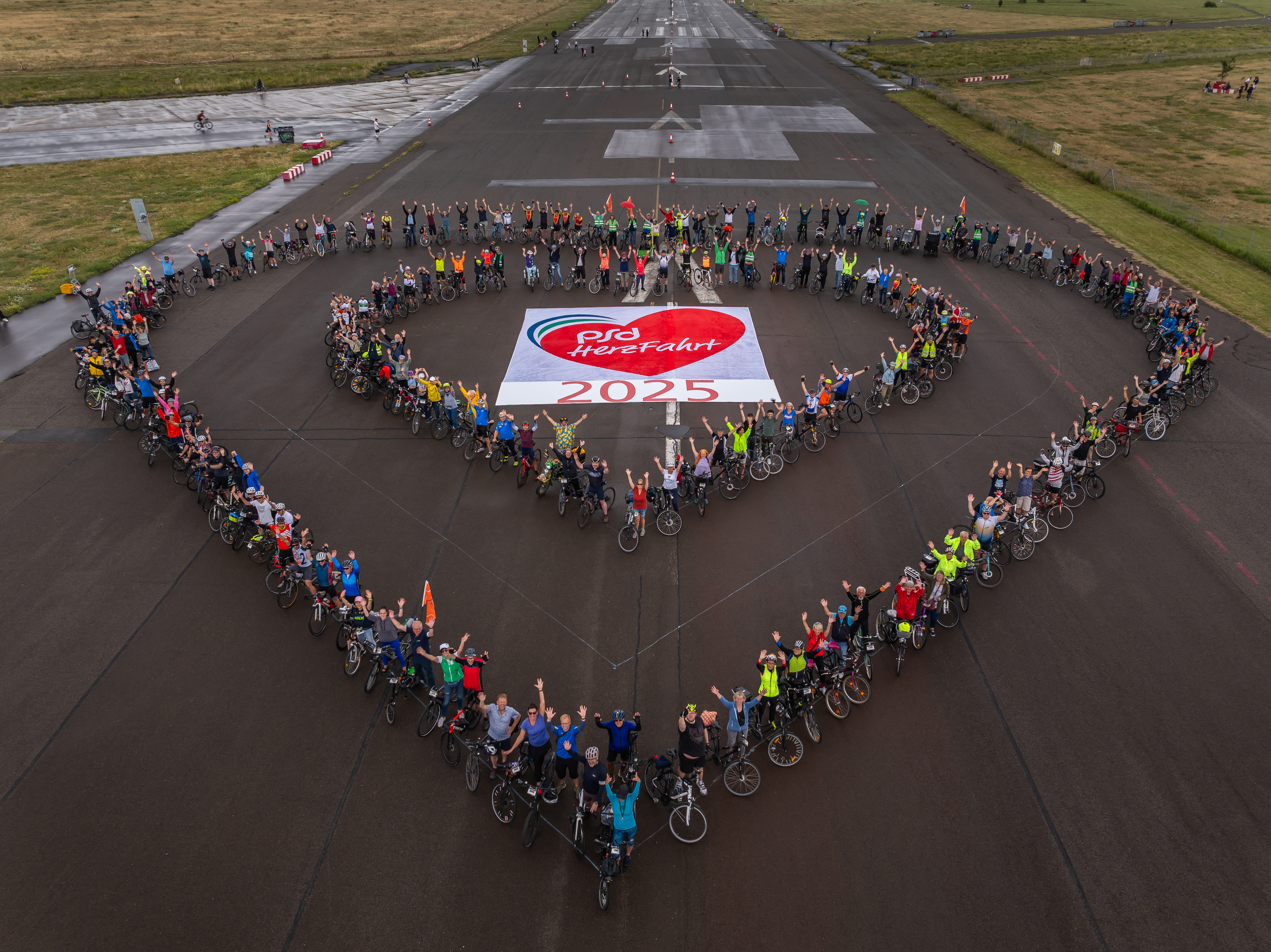 Eine Gruppe von Radfahrern steht auf einem Flugplatz und bildet gemeinsam ein Herz. In der Mitte liegt ein Banner mit der Aufschrift "PSG HereFahrt 2025". Viele Teilnehmer heben die Hände und sind in bunten Outfits gekleidet. Es herrscht eine fröhliche Atmosphäre.