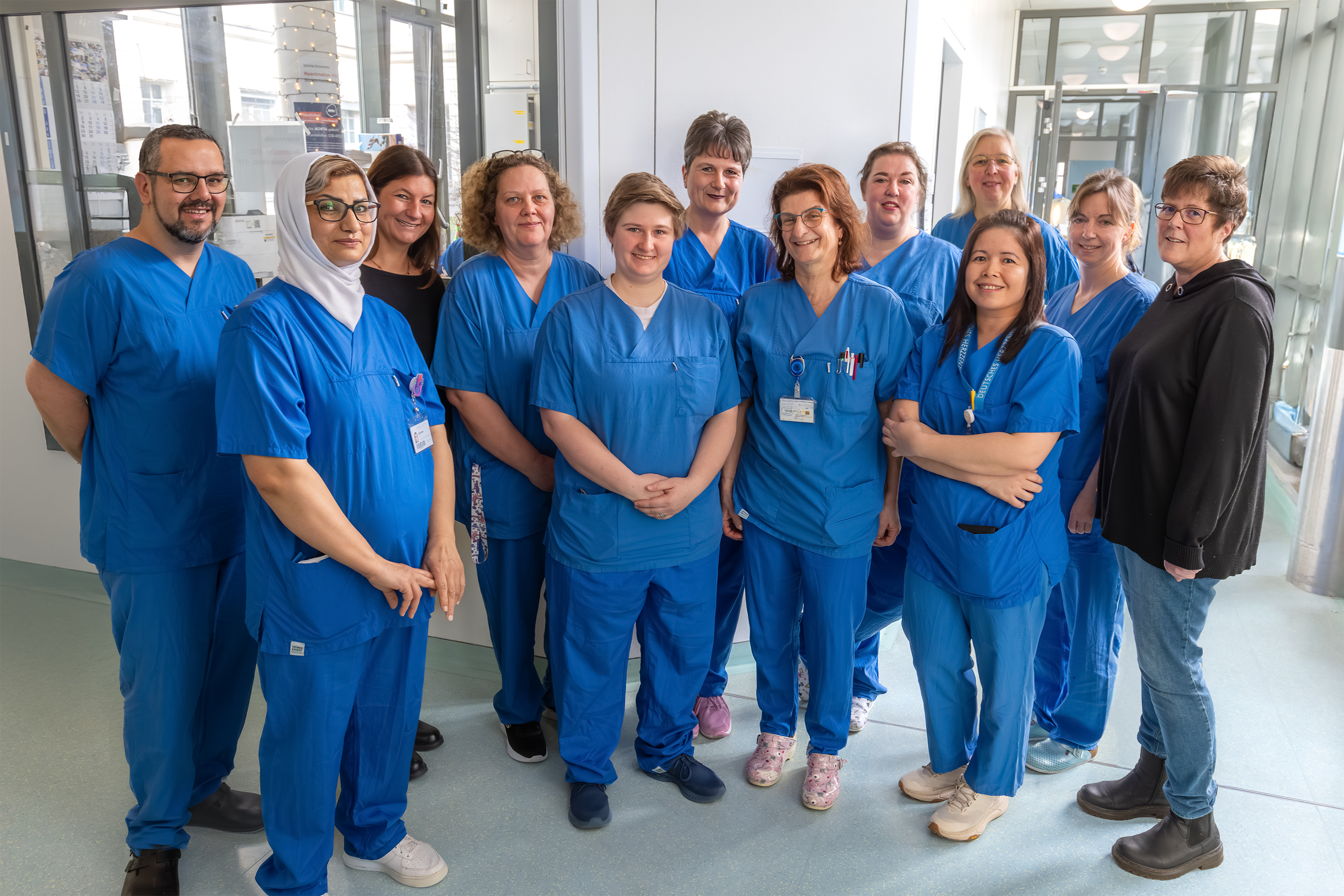 Eine Gruppe von 15 Personen in blauen Medizin-Overalls steht in einem hellen Raum. Sie lächeln und posieren miteinander. Hinter ihnen sind Fenster und ein heller Boden sichtbar, was auf eine medizinische Einrichtung hinweist.