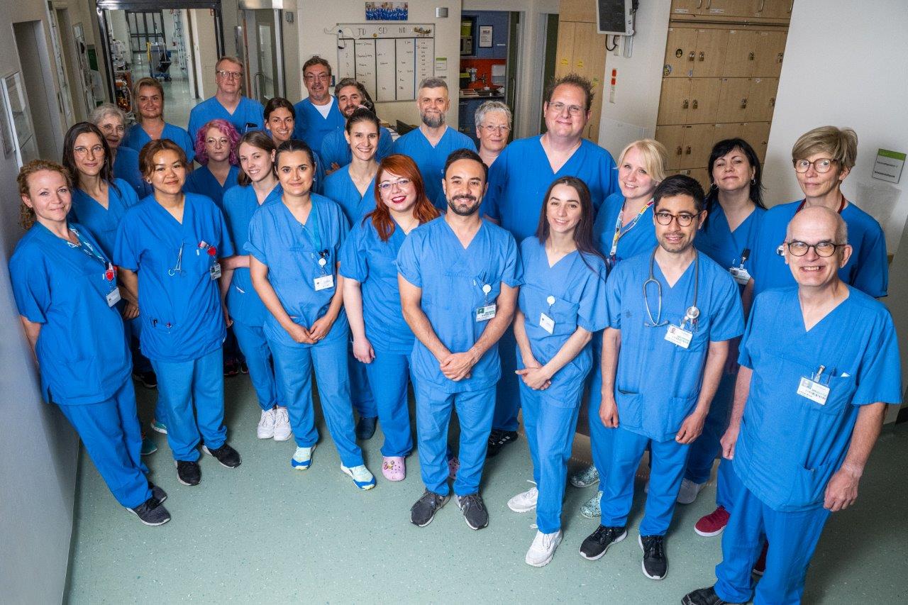 Eine Gruppe von etwa 25 Personen in blauen Krankenhausuniformen steht gemeinsam in einem Flur. Sie lächeln und wirken freundlich. Im Hintergrund sind medizinische Utensilien und Schränke sichtbar, die auf eine gesundheitliche Einrichtung hinweisen.