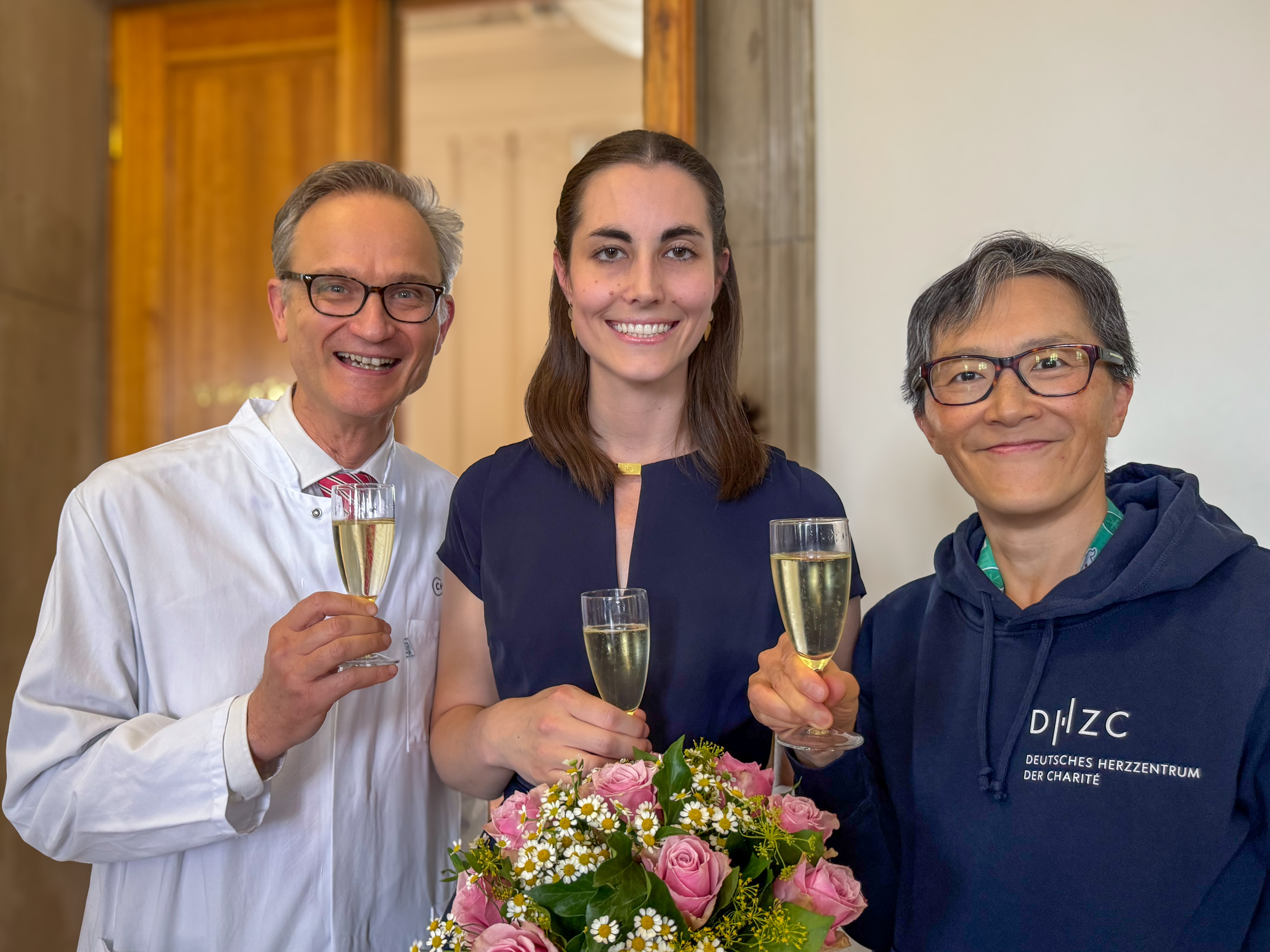 Drei Personen stehen zusammen und stoßen mit Sektgläsern an. Sie lächeln und präsentieren einen Blumenstrauß mit rosa Rosen und weißen Blumen im Vordergrund. Die Szene vermittelt eine festliche Stimmung.