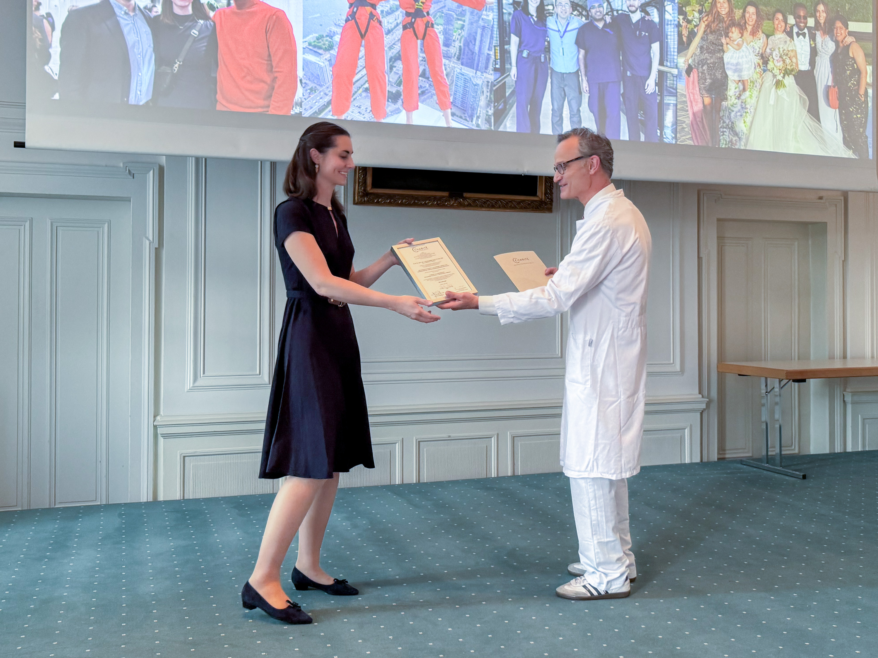 Eine Frau in einem schwarzen Kleid steht auf einem Teppich und nimmt ein Dokument von einem Mann in einem weißen Laborkittel entgegen. Beide lächeln, während sie sich gegenüberstehen. Im Hintergrund ist eine große Präsentationsleinwand zu sehen.