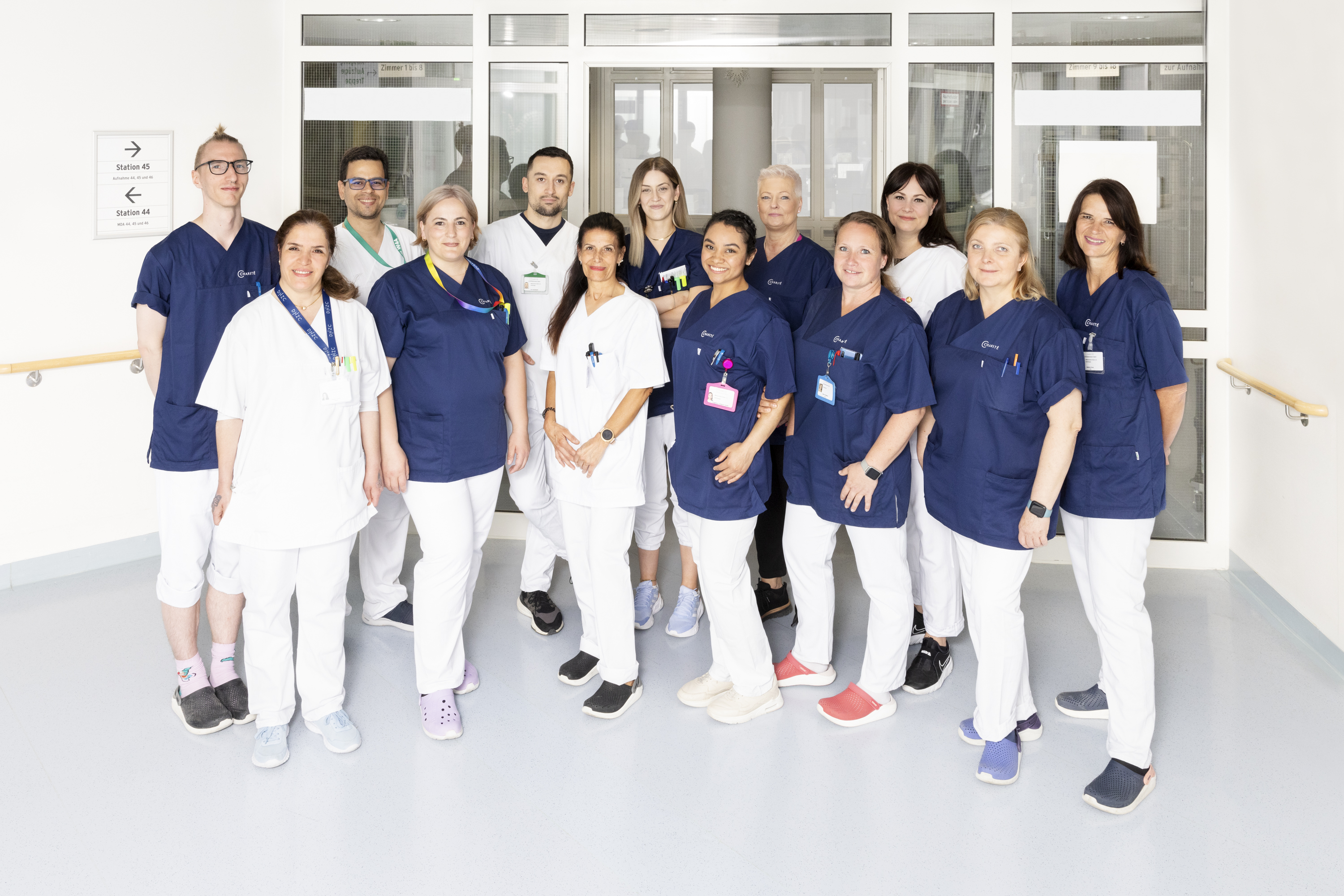 Eine Gruppe von 14 Personen in medizinischer Kleidung steht in einem Krankenhausflur. Die meisten tragen blaue Oberteile und weiße Hosen. Sie lächeln und stehen in einer lockeren Formation, wobei einige Namensschilder und Lanyards sichtbar sind. Im Hintergrund sind Glastüren zu sehen.