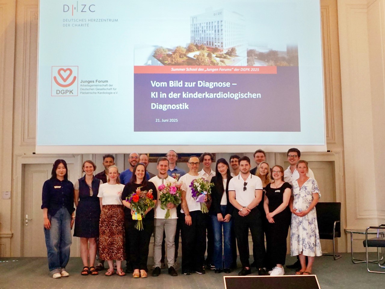 Eine Gruppe von 18 Personen steht im Vordergrund und hält Blumen. Im Hintergrund befindet sich eine Projektionsfläche mit dem Titel „Vom Bild zur Diagnose – KI in der kinder-kardiologischen Diagnostik“ und dem Datum 21. Juni 2025.