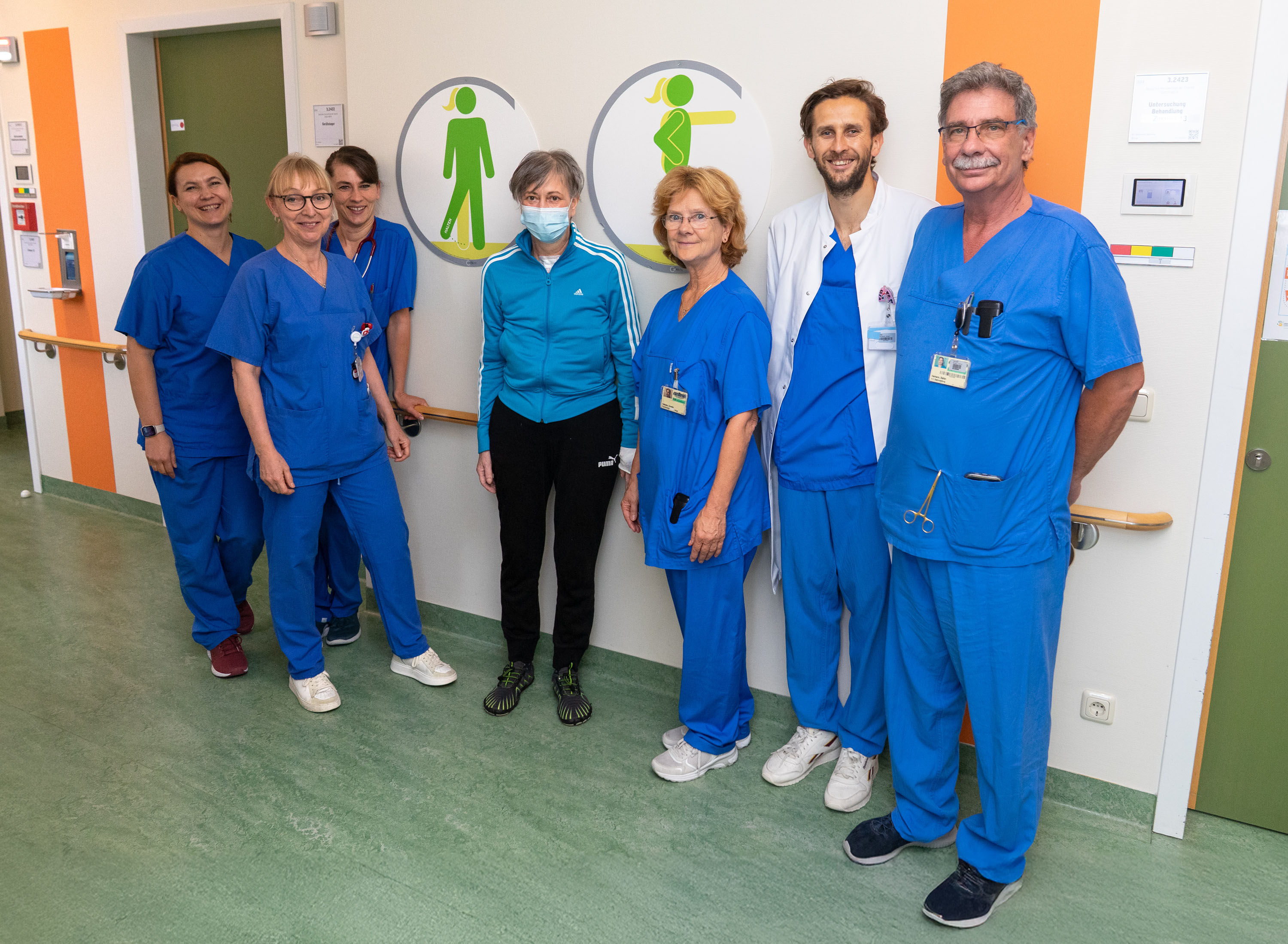 Eine Gruppe von acht Personen steht in einem Krankenhausflur. Die meisten tragen medizinische Kleidung in Blau, während eine Frau in der Mitte eine Maske trägt und Sportbekleidung anhat. Hinter ihnen sind Symbole für Männer und Frauen auf der Wand zu sehen.