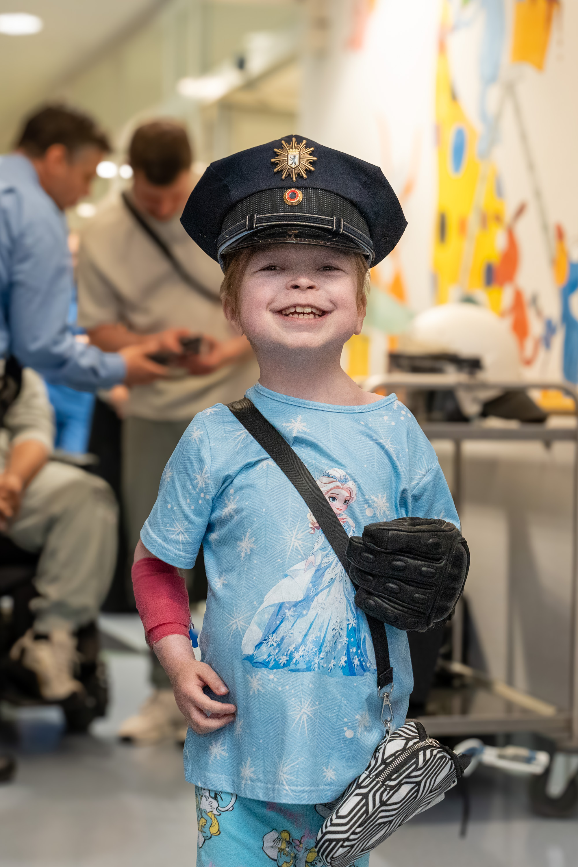 Ein lächelndes Kind trägt eine Polizeikappe und ein T-Shirt mit einem Eisprinzessinnen-Motiv. Es hat eine kleine Tasche über der Schulter und steht fröhlich im Vordergrund, während im Hintergrund andere Menschen beschäftigt sind.