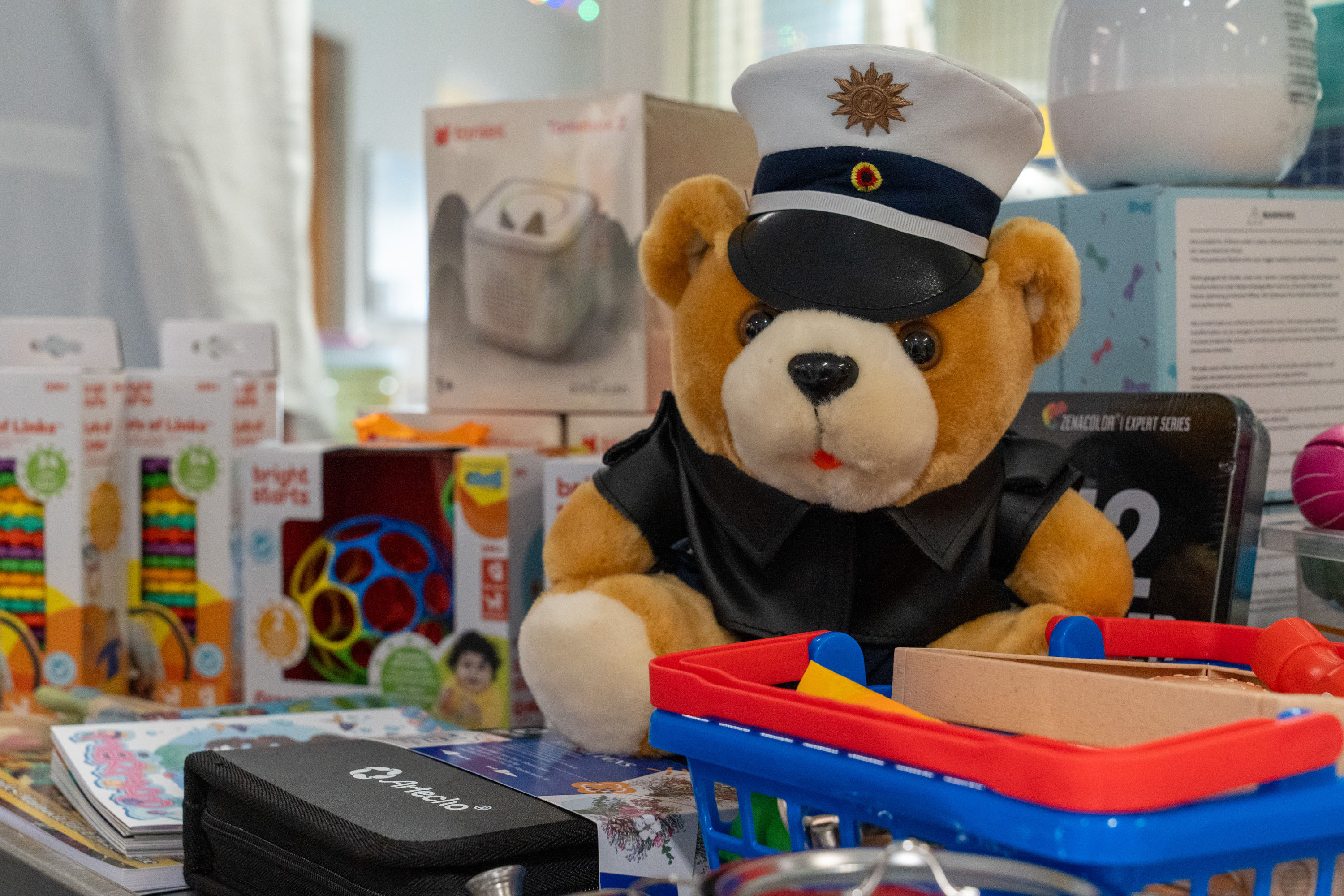 Ein Plüschbär in einer Polizeiuniform sitzt auf einem Tisch, umgeben von Spielzeugen und Bastelmaterialien. Der Bär trägt eine Mütze mit einer Abzeichen und hat einen freundlichen Gesichtsausdruck. Im Vordergrund steht ein kleiner Einkaufskorb mit verschiedenen Spielzeugen.