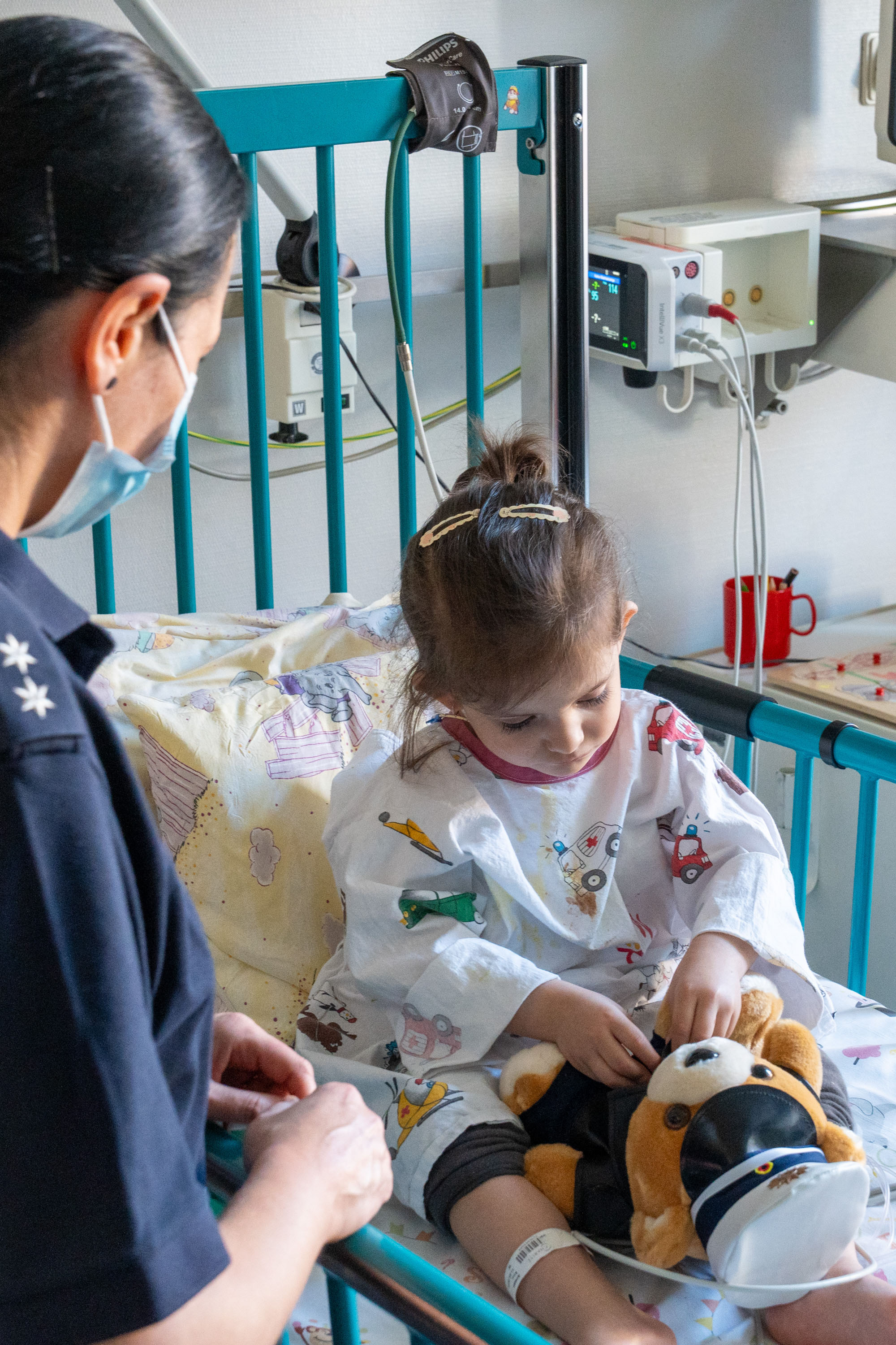 Ein kleines Mädchen sitzt in einem Krankenhausbett und spielt mit einem Stofftier. Eine Krankenschwester steht neben ihr und beobachtet sie. Das Mädchen trägt ein Krankenhaushemd und hat einen Verband am Arm. Die Atmosphäre ist freundlich und fürsorglich.