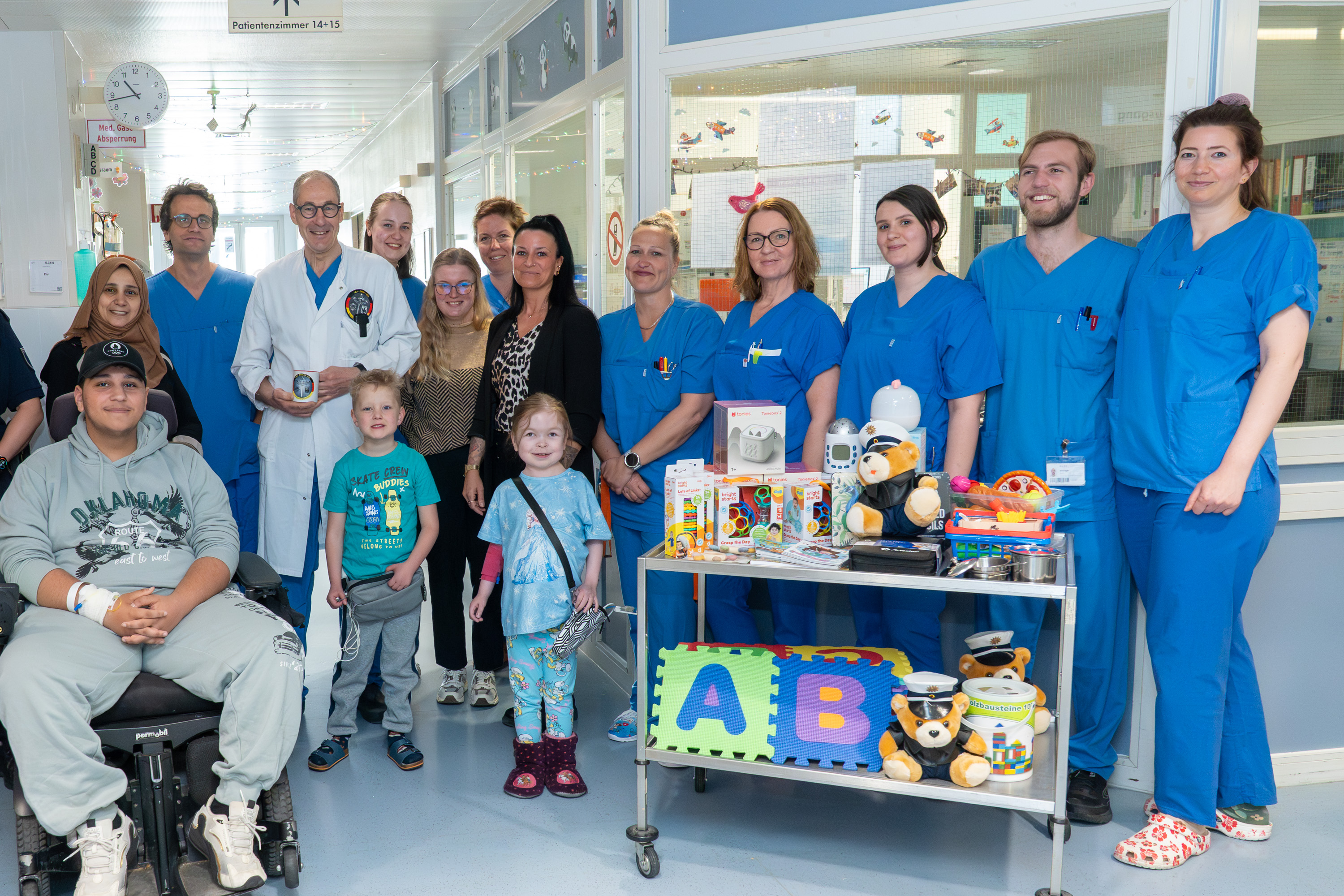 Eine Gruppe von Menschen, darunter Patienten und medizinisches Personal, steht in einem Krankenhausflur. Einige tragen blaue Arbeitskleidung, und es gibt einen Rollstuhlfahrer. Vor ihnen steht ein Wagen mit Spielsachen und Lernmaterialien, der zur Unterstützung von Kindern dient.