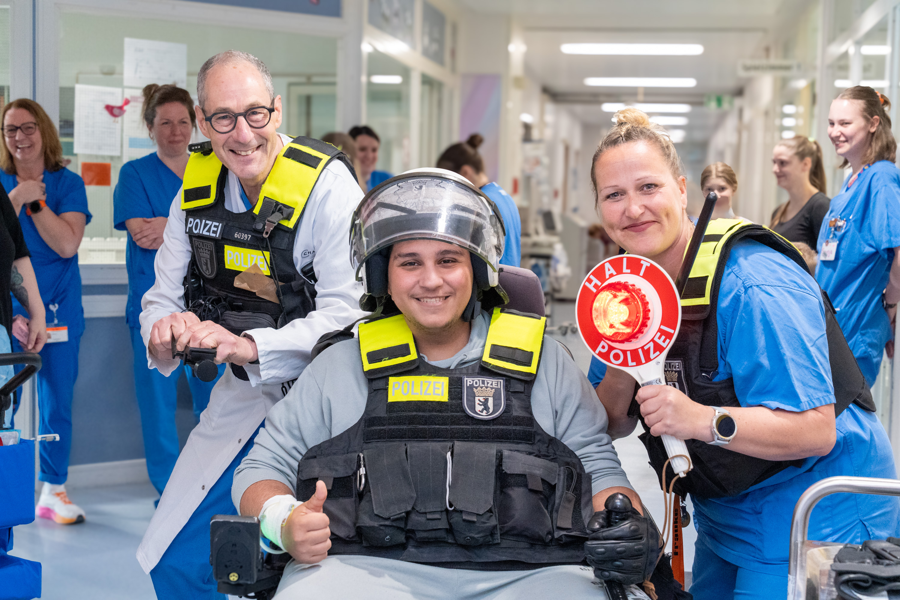 Eine Gruppe von Menschen in einem Flur eines Krankenhauses. Ein Mann sitzt lächelnd in einem Rollstuhl, trägt Schutzkleidung und ein Helm. Neben ihm stehen zwei Personen in Uniform, die ebenfalls lächeln. Im Hintergrund sieht man medizinisches Personal in blauen Kleidern.