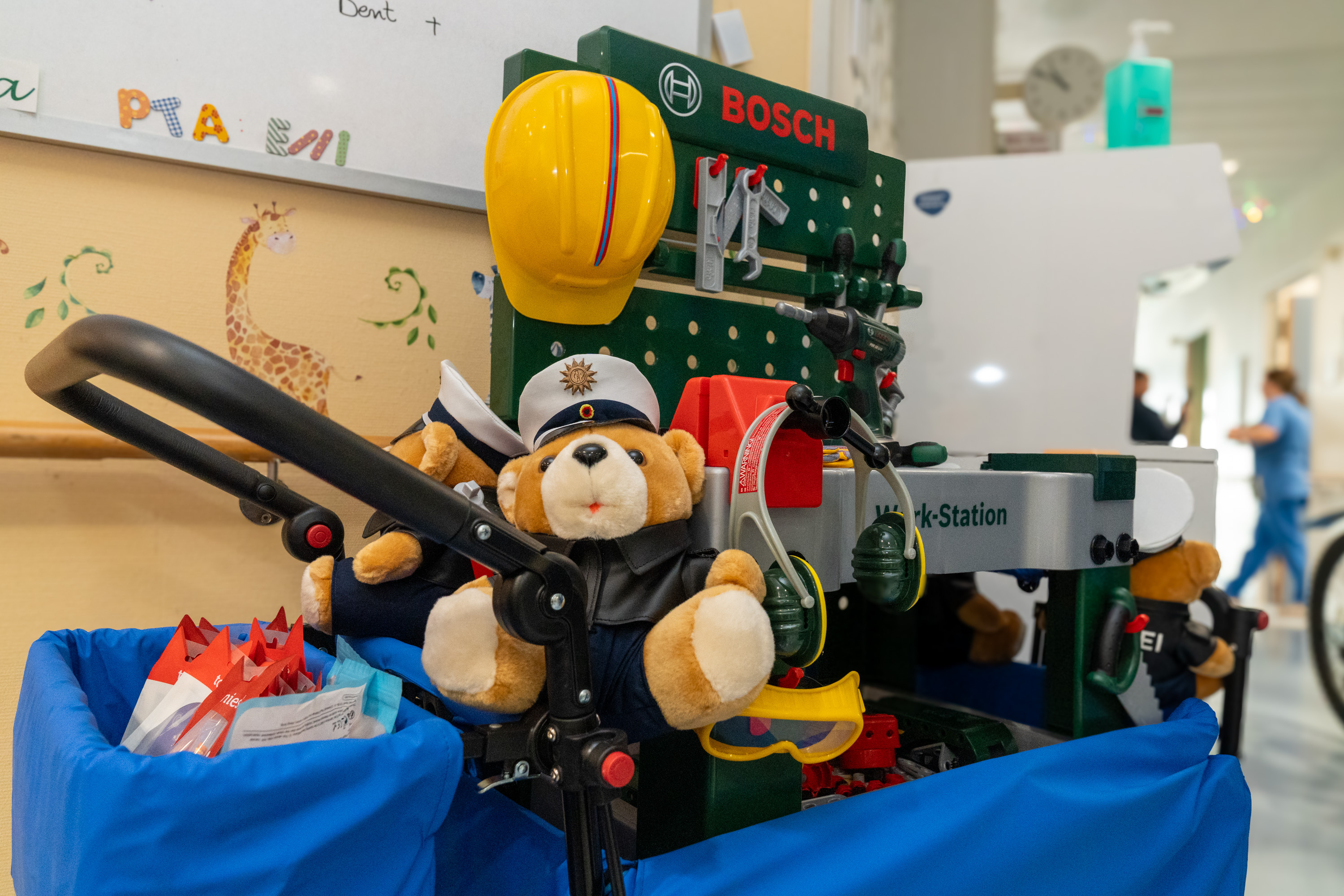 Ein Wagen mit Spielzeugcontainern, auf dem sich ein brauner Teddybär in einer Uniform befindet. Der Bär trägt einen Hut und ist von Werkzeugen sowie einem gelben Helm umgeben. Der Hintergrund zeigt eine kindgerechte Umgebung in einem spielerischen Kontext.