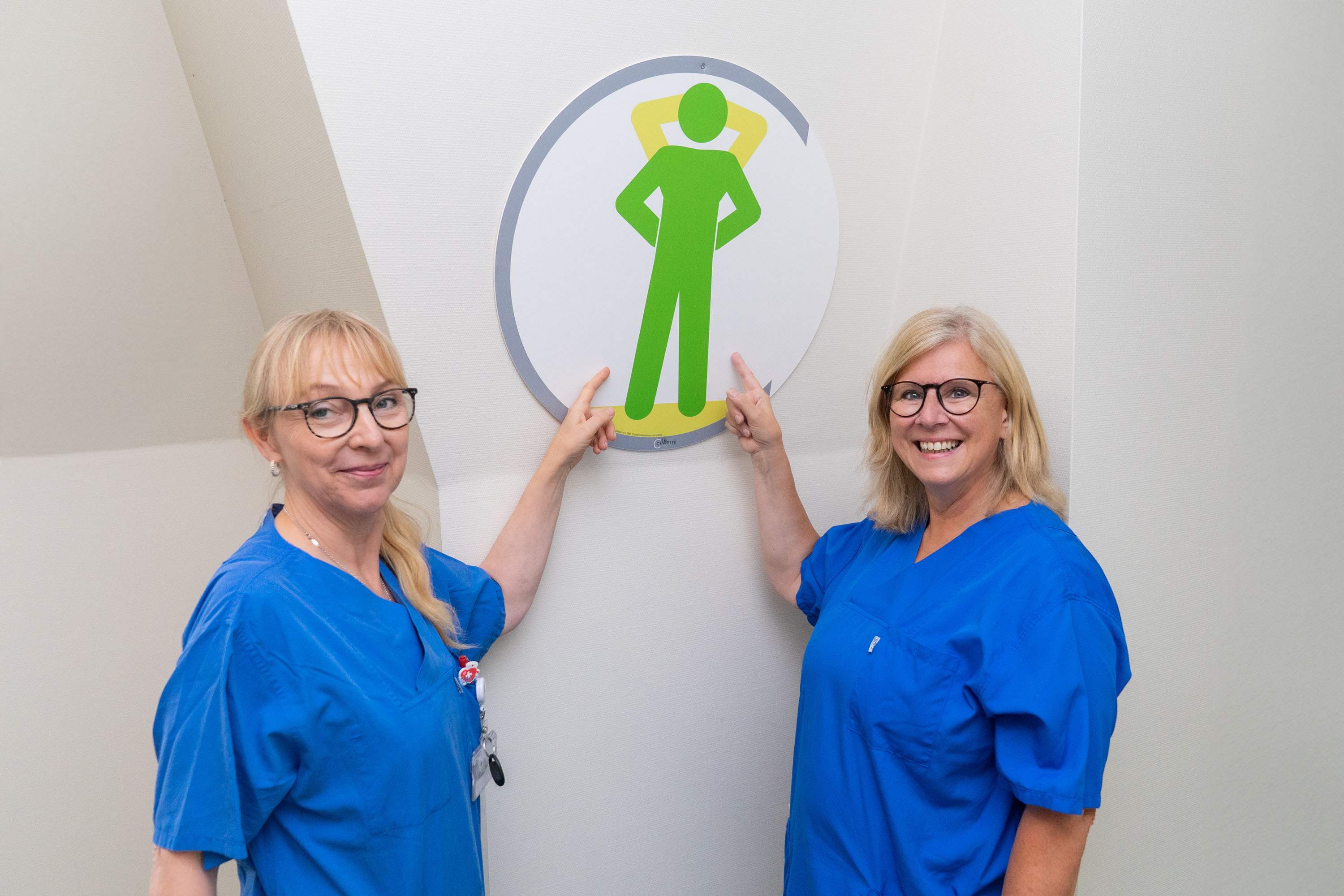 Zwei Frauen in blauen Kitteln stehen vor einer Wand. Sie zeigen auf ein großes, rundes Schild mit einer grünen, stilisierten Figur, die mit erhobenen Armen dargestellt ist. Die Frauen lächeln freundlich in die Kamera.