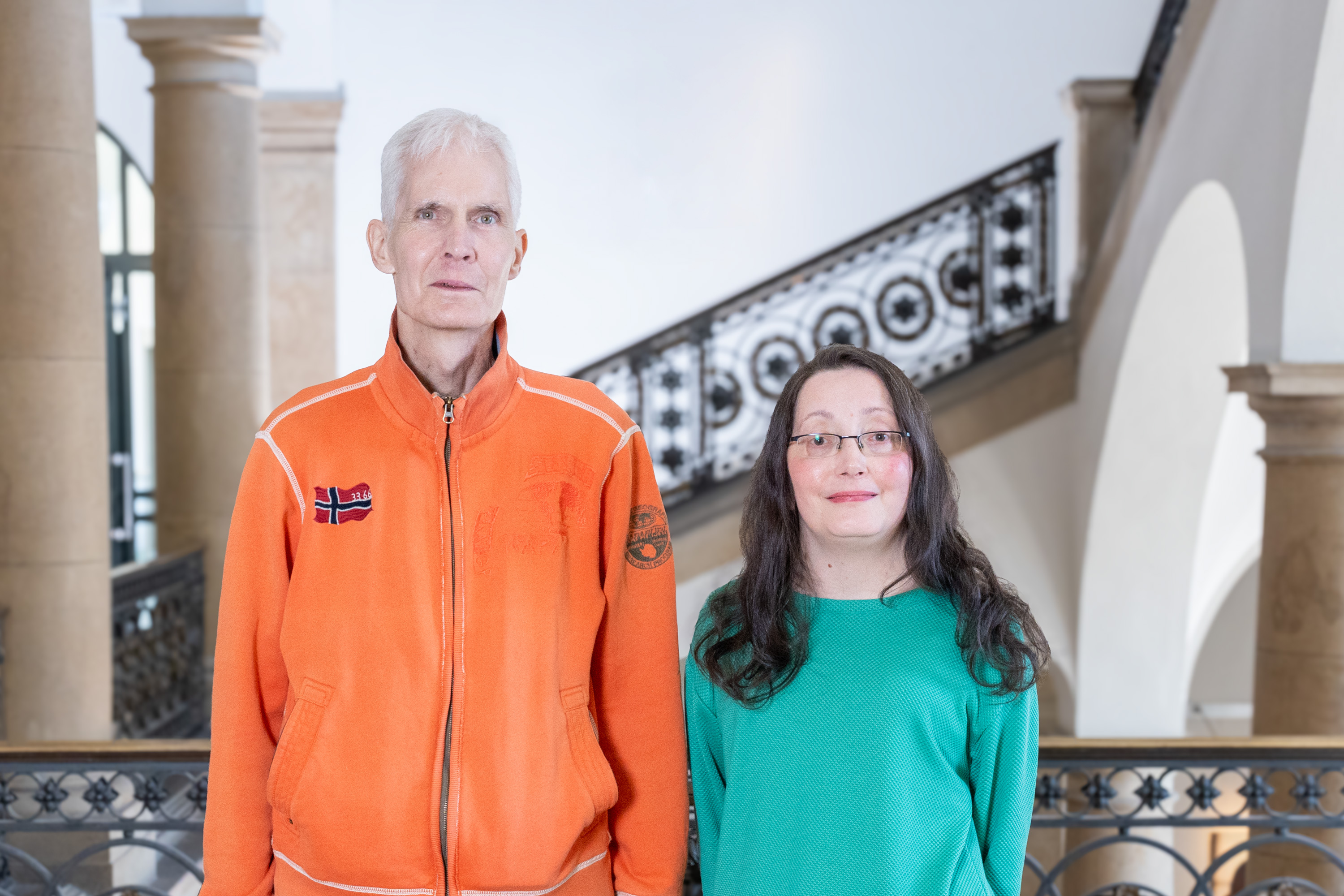 Das Bild zeigt einen Mann in einer orangefarbenen Jacke und eine Frau in einem grünen Oberteil. Sie stehen nebeneinander in einem Gebäude mit einem eleganten Geländer und Stufen im Hintergrund. Beide blicken direkt in die Kamera.