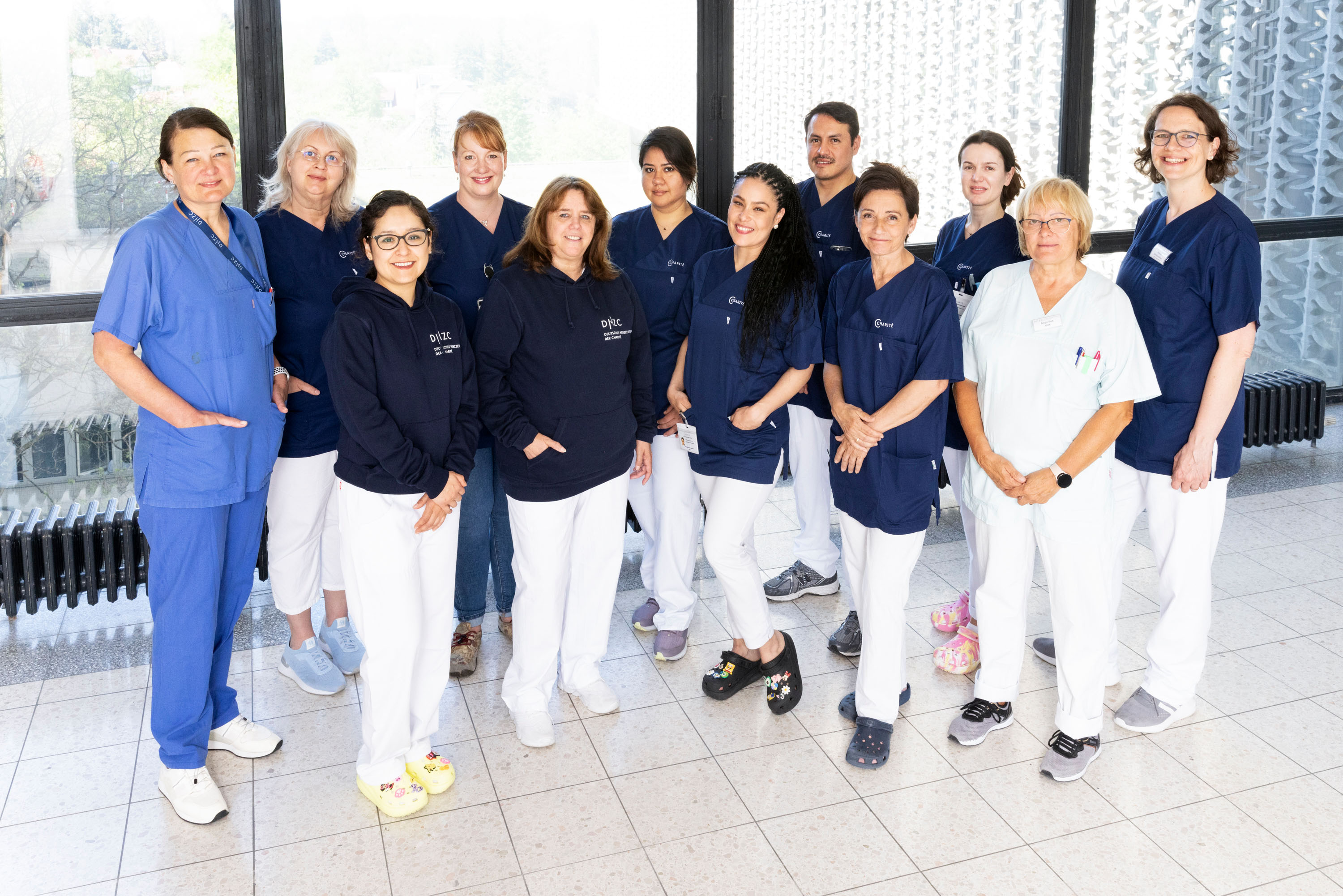 Eine Gruppe von 14 Personen steht in einem hellen Raum. Sie tragen hauptsächlich dunkelblaue und weiße Arbeitskleidung. Die Mitglieder des Teams lächeln und zeigen eine freundliche Haltung. Im Hintergrund sind große Fenster, durch die Licht strahlt.