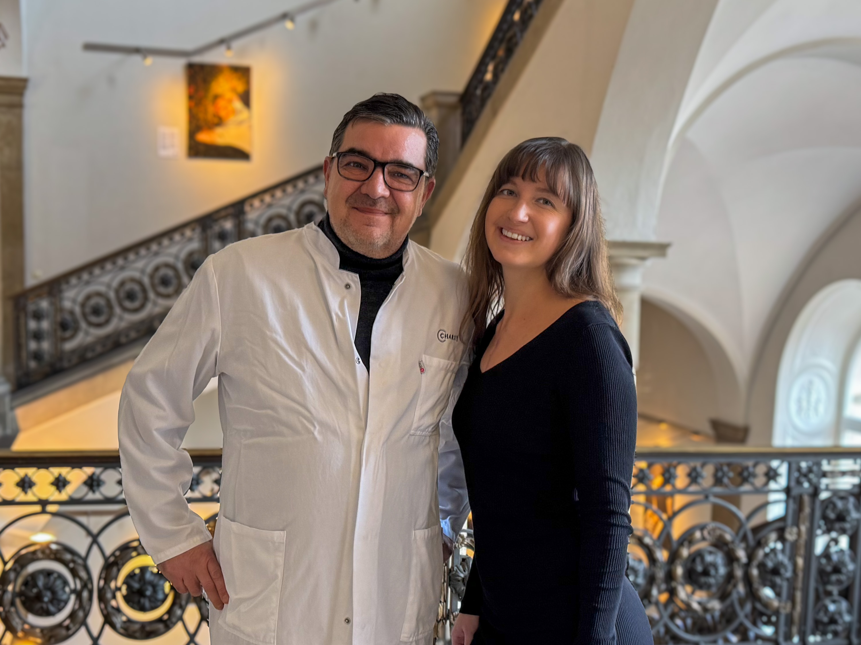 Zwei Personen stehen vor einem geschwungenen Geländer in einem eleganten Innenraum. Der Mann trägt einen weißen Laborkittel und Brille, die Frau trägt ein schwarzes Oberteil und lächelt. Im Hintergrund ist eine Treppe und ein Kunstwerk an der Wand sichtbar.