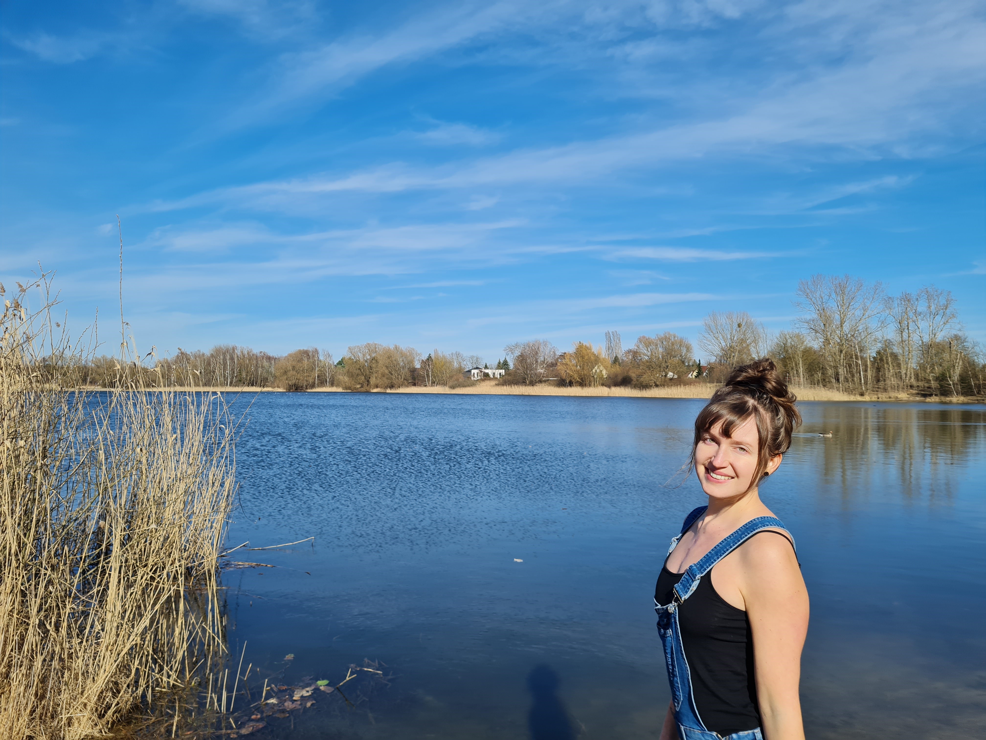 Eine junge Frau in einem schwarzen Top und Latzhose lächelt am Ufer eines ruhigen Sees. Im Hintergrund sind Bäume und ein klarer Himmel zu sehen. Die Szenerie strahlt eine freundliche und entspannte Atmosphäre aus.