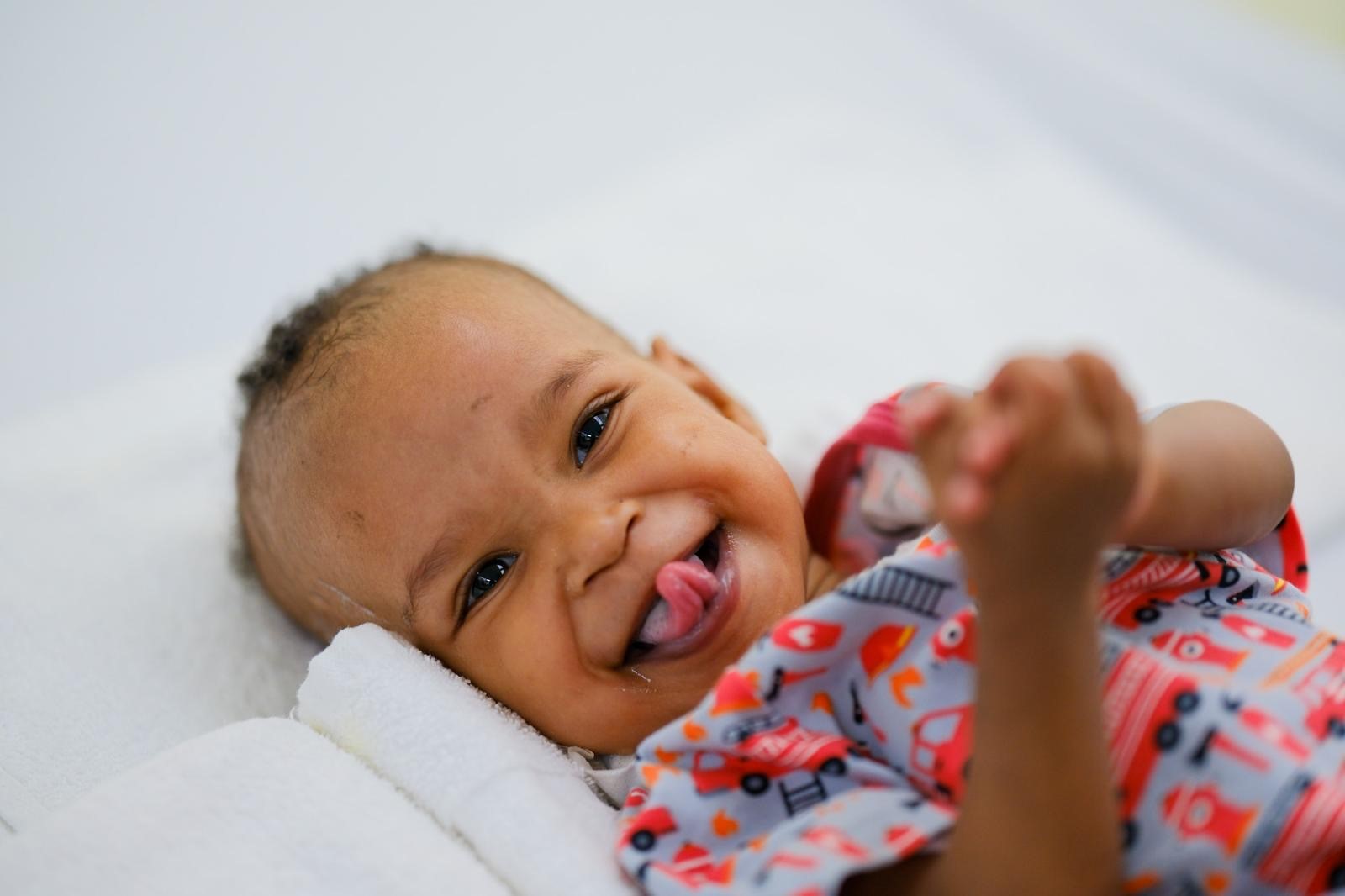 Ein lächelndes Baby liegt auf einer Liege, trägt ein buntes Oberteil mit Fahrzeugmotiven und zeigt die Zunge. Die Augen sind lebhaft, und die Hände sind zusammengelegt, als ob es sich freut. Der Hintergrund ist unscharf und hell.