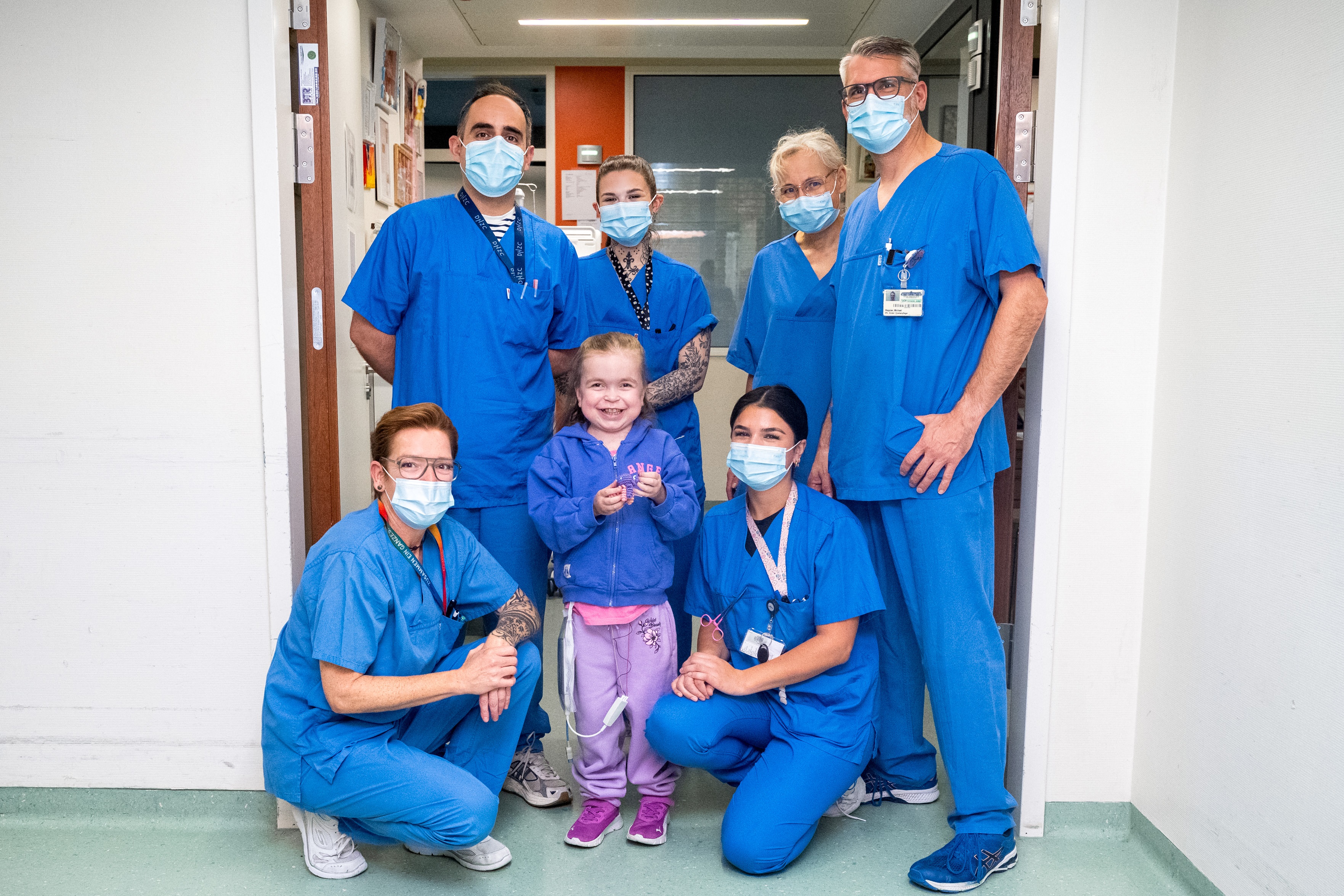 Eine Gruppe von sechs Klinikmitarbeitern in blauen Scrubs und Schutzmasken steht um ein kleines Mädchen, das ein lächelndes Gesicht zeigt und eine Puppe in der Hand hält. Die Mitarbeiter wirken freundlich und unterstützend, um die positive Atmosphäre im Gesundheitswesen zu betonen.