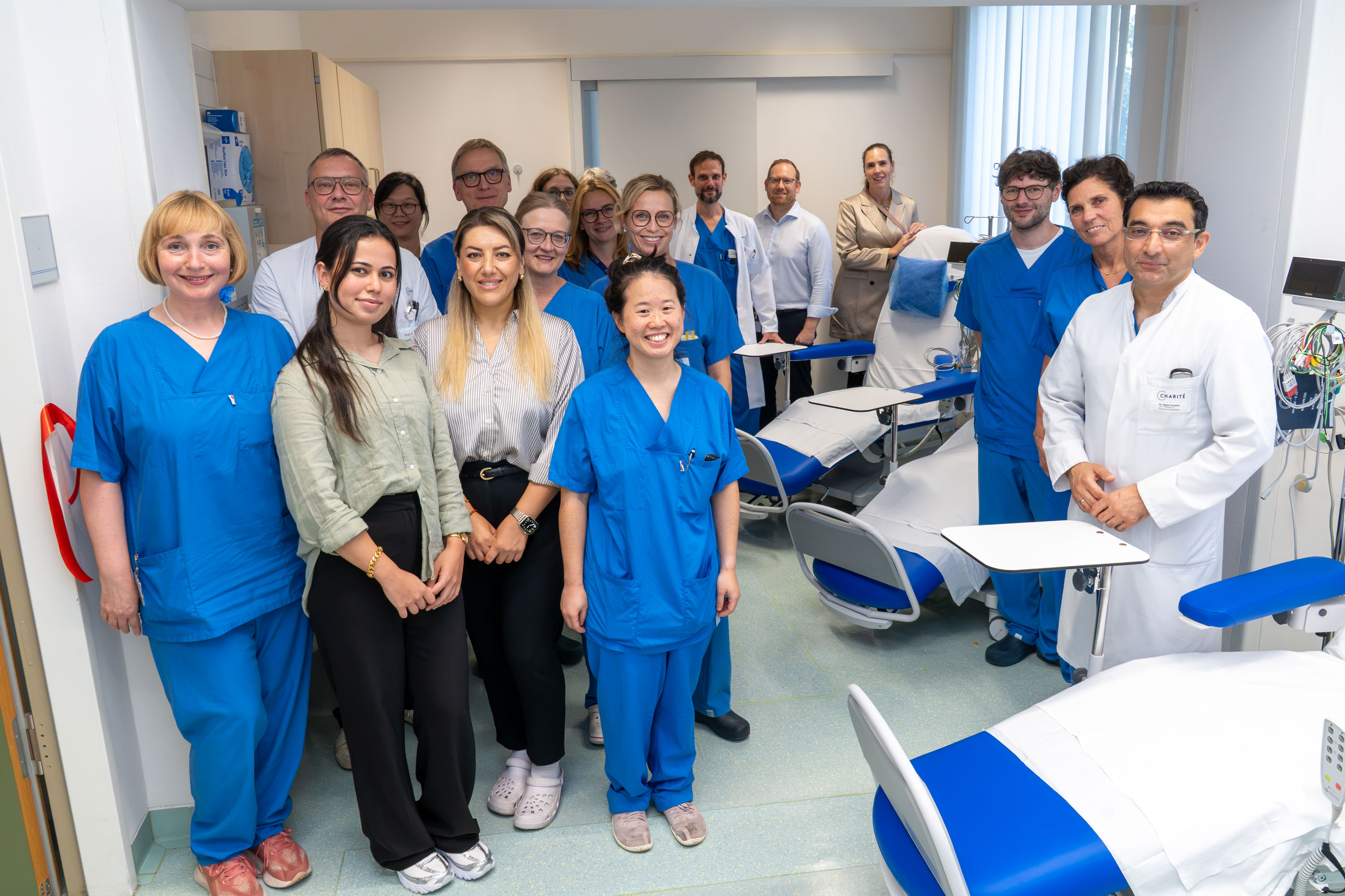 Eine Gruppe von 15 Personen steht in einer Klinik, gekleidet in blaue Arbeitskleidung. Im Hintergrund sind medizinische Geräte und Untersuchungsstühle zu sehen. Die Atmosphäre wirkt freundlich und kollegial. Einige Personen lächeln in die Kamera.