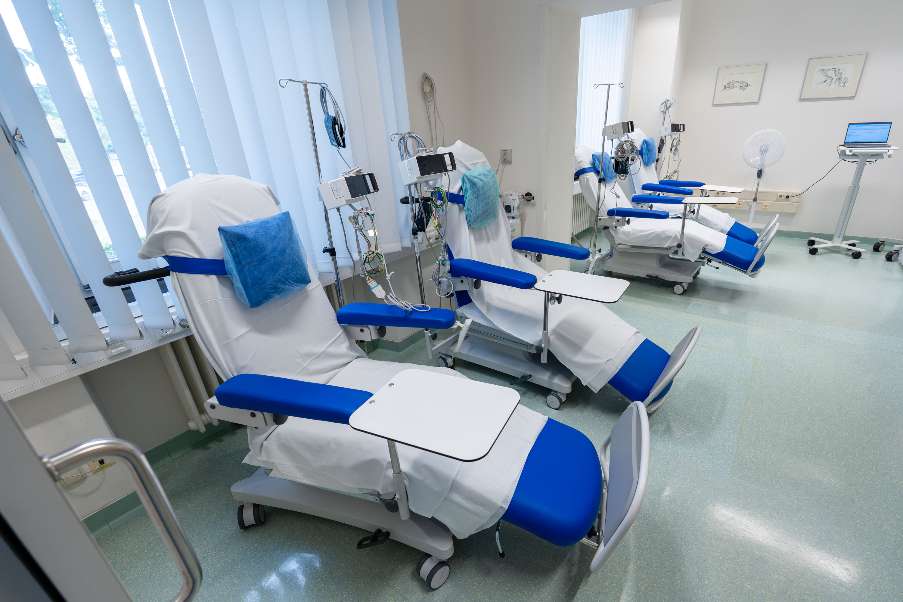 Ein heller Raum mit mehreren medizinischen Behandlungsstühlen. Die Stühle sind mit blauen und weißen Abdeckungen versehen und verfügen über kleine Ablagetische. Im Hintergrund sind medizinische Geräte und ein Monitor sichtbar. Die Fenster sind mit hellen Vorhängen versehen.