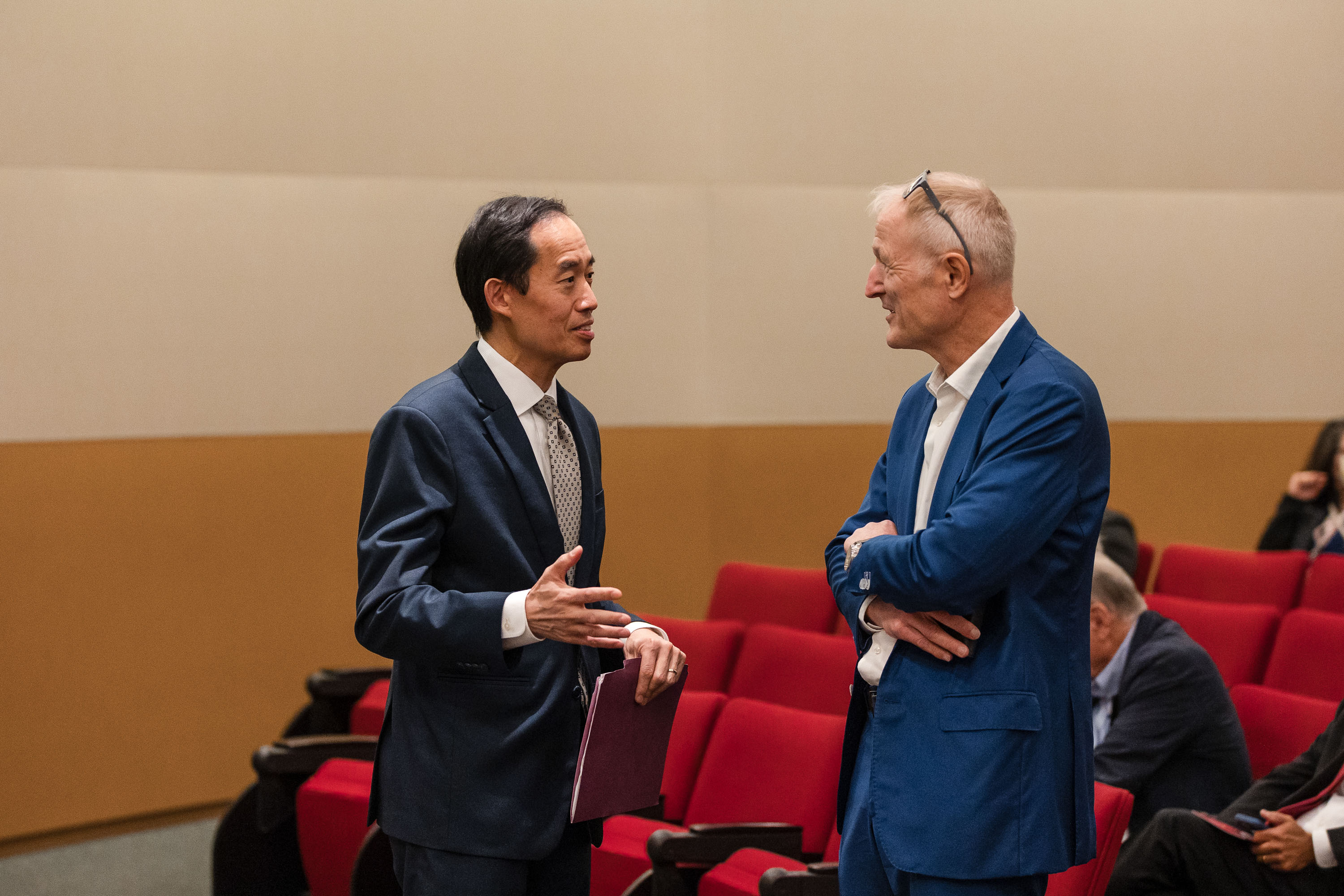 Zwei Männer in Anzügen stehen in einem Konferenzraum und unterhalten sich. Im Hintergrund sind leere rote Sitzreihen sichtbar. Einer der Männer trägt eine Sonnenbrille auf dem Kopf. Die Atmosphäre wirkt informell und freundlich.