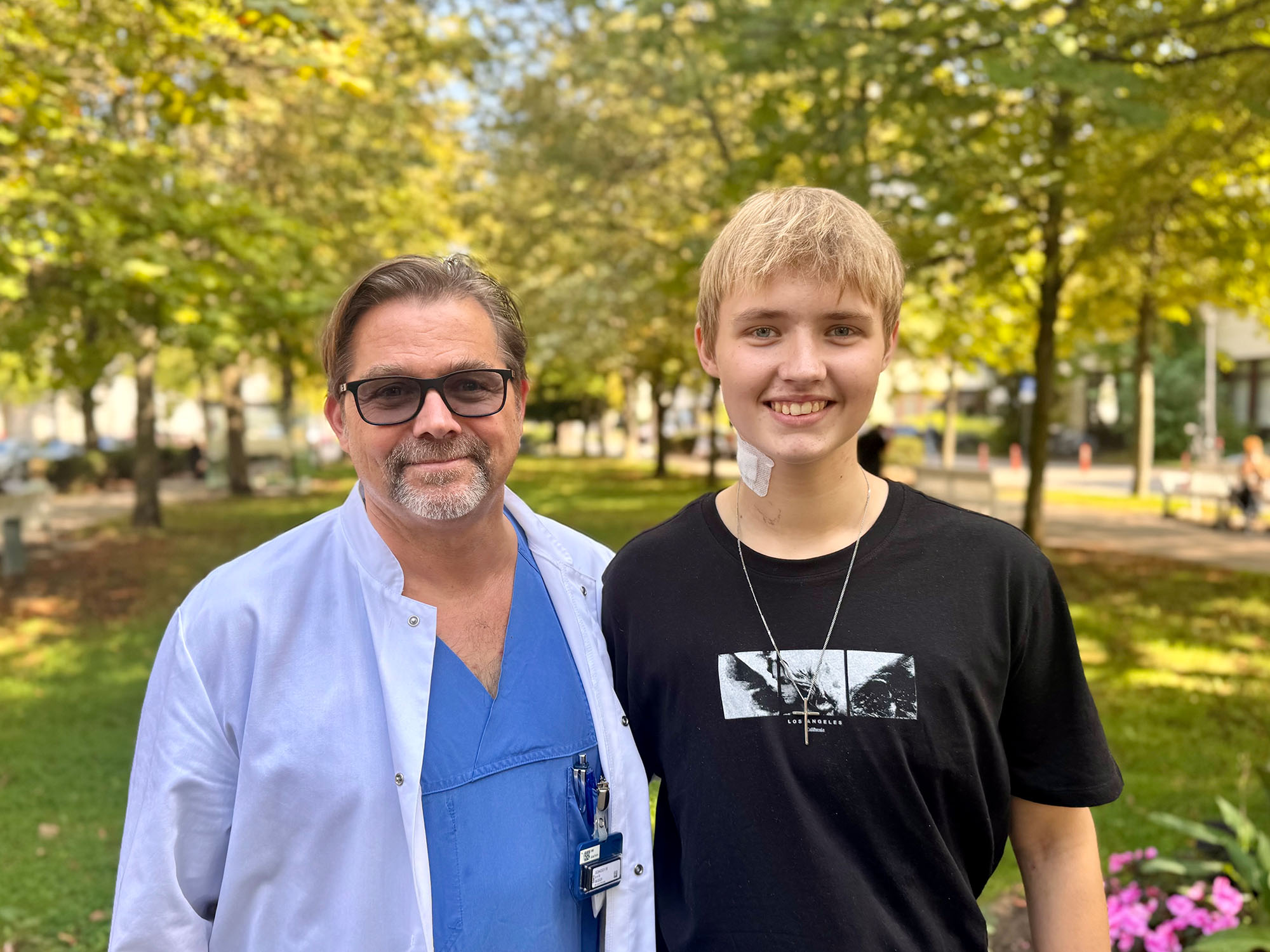 Ein männlicher Arzt mit Brille in medizinischer Kleidung steht neben einem jungen Mann, der ein schwarzes T-Shirt trägt. Beide lächeln und befinden sich in einem Park mit Bäumen und Blumen im Hintergrund.