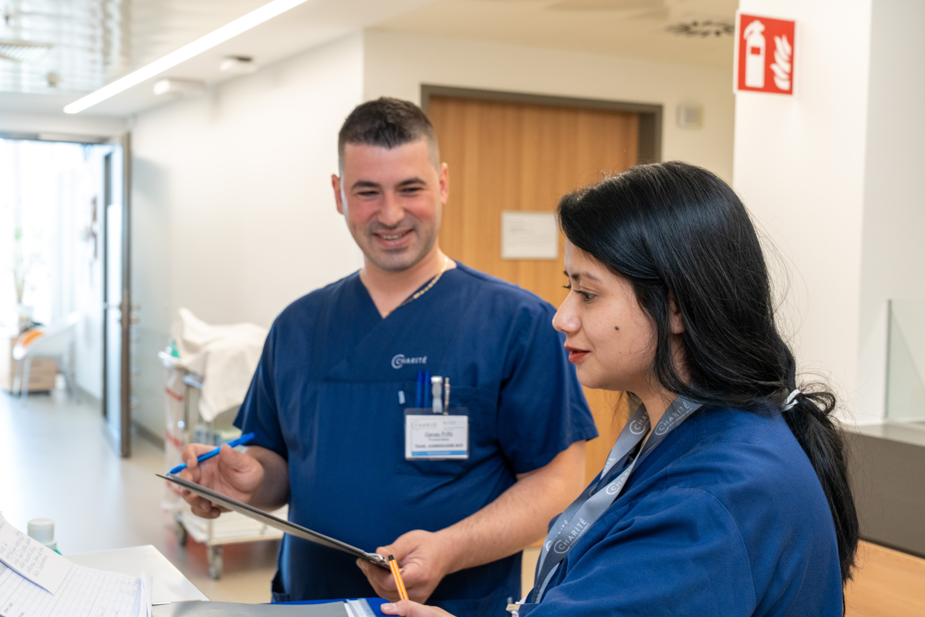 Zwei Pflegekräfte in blauen Uniformen stehen an einem Empfangstresen. Sie schauen sich Unterlagen an und diskutieren. Der Mann lächelt, während die Frau aufmerksam zuhört. Im Hintergrund sind Krankenhausmöbel und eine Tür sichtbar.