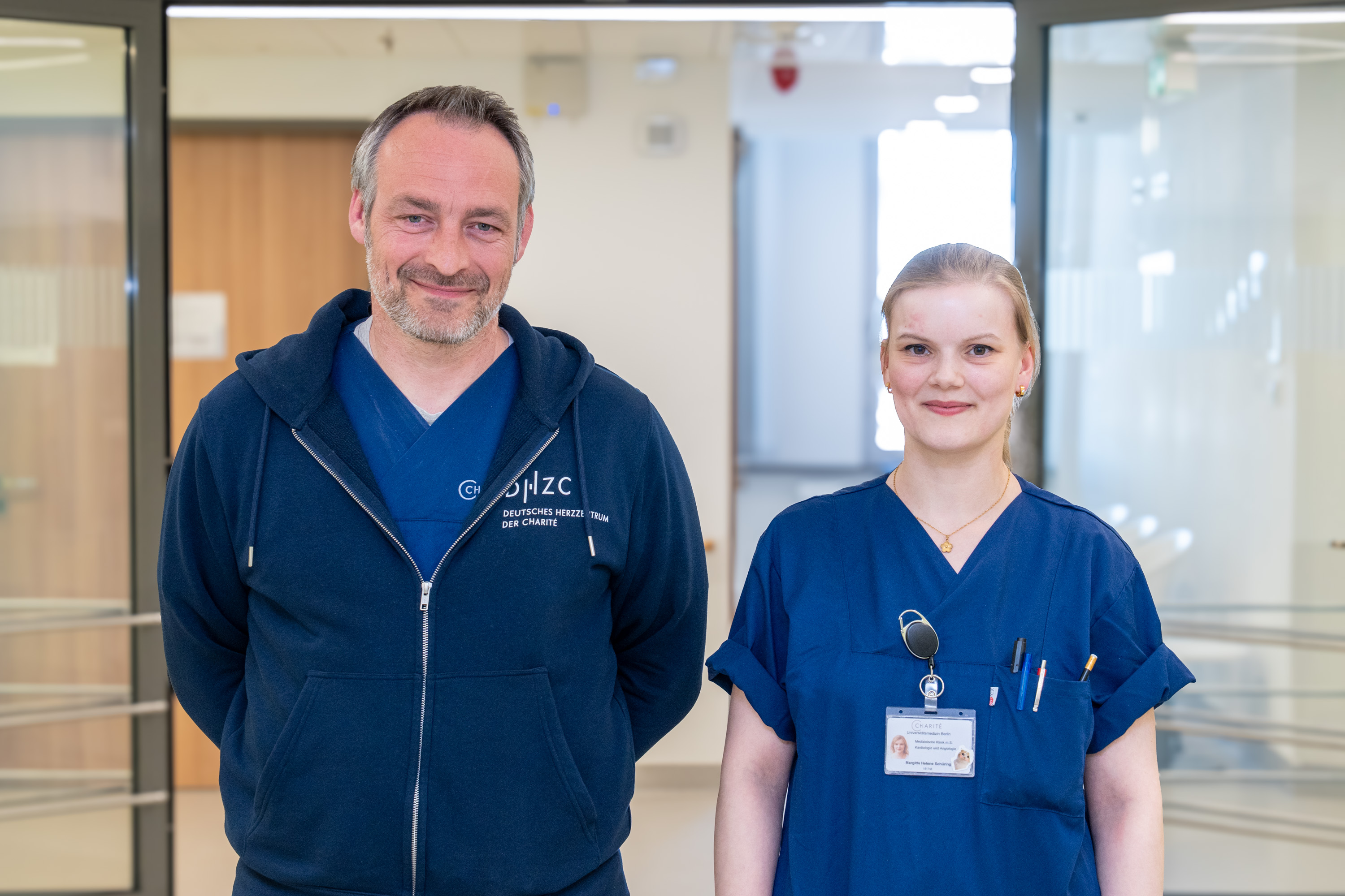 Zwei medizinische Fachkräfte stehen in einem Krankenhausflur. Der Mann trägt einen blauen Hoodie und ein T-Shirt, die Frau ein dunkelblaues Oberteil mit Namensschild und Stethoskop. Beide lächeln freundlich und scheinen bereit, Hilfe zu leisten.