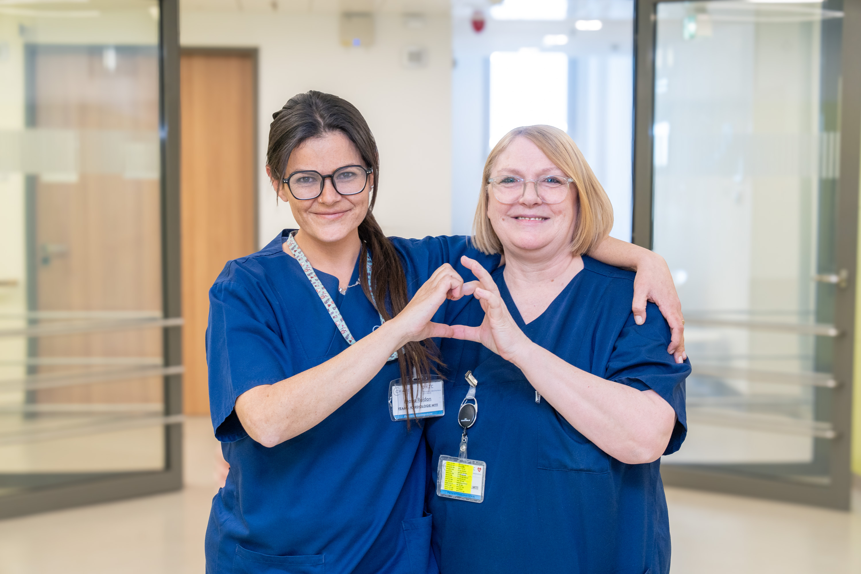 Zwei Frauen in blauen medizinischen Kitteln stehen in einem modernen Gesundheitsumfeld. Sie formen mit ihren Händen ein Herz und lächeln, was Zuneigung und Teamarbeit symbolisiert.