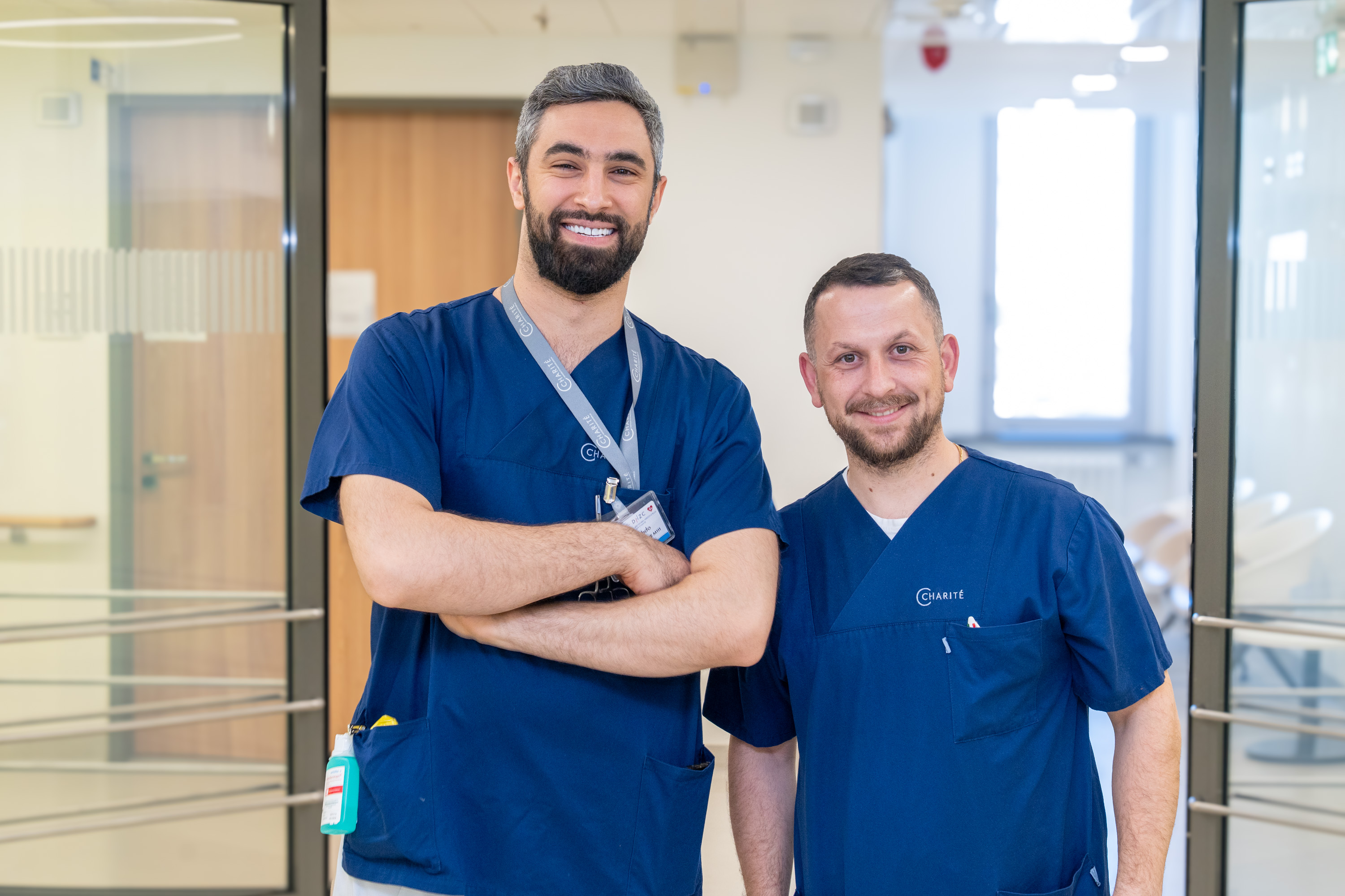 Zwei Männer in blauer Arbeitskleidung stehen in einem Krankenhausflur. Der Mann links hat einen kurzen Bart und lächelt, während der Mann rechts ebenfalls lächelt. Beide wirken freundlich und entspannt. Im Hintergrund sind Fenster und helle Wände sichtbar.