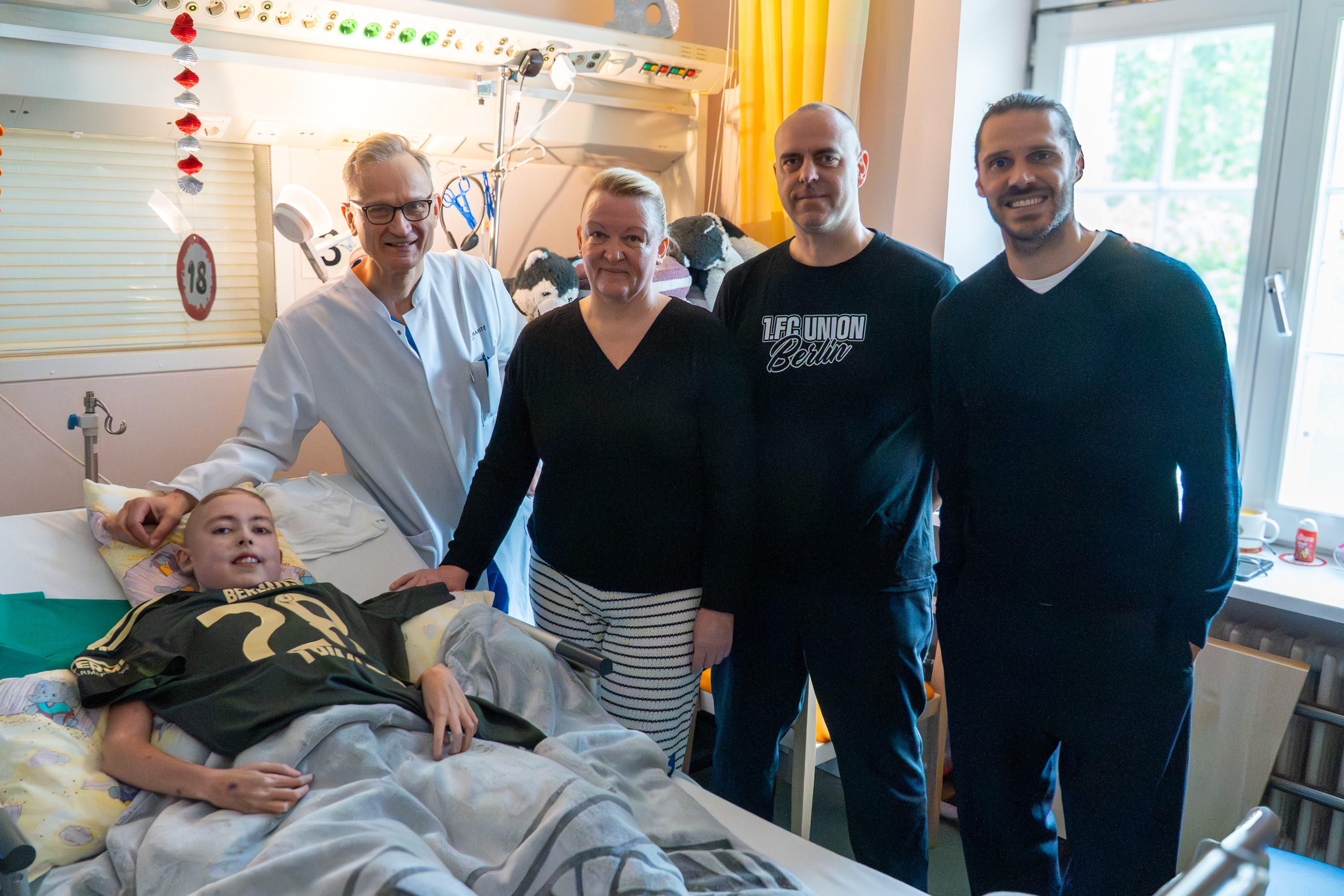 Eine Gruppe von fünf Personen steht um ein krankes Kind, das im Krankenhausbett liegt. Der Junge trägt ein grünes T-Shirt und schaut lächelnd. Die Erwachsenen stehen eng beieinander und zeigen eine unterstützende Haltung.