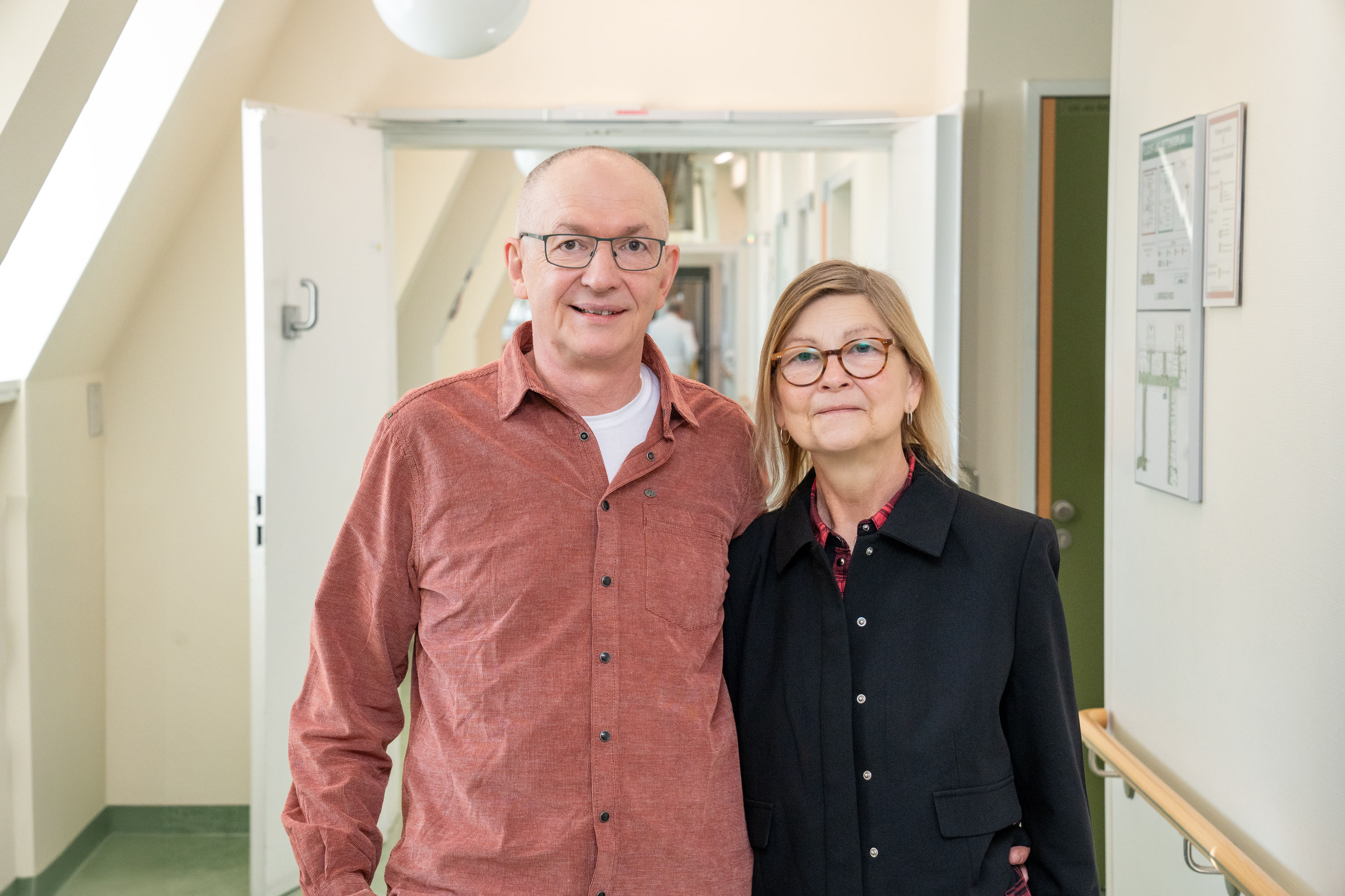 Ein älteres Paar steht lächelnd in einem Flur. Der Mann trägt ein rotes Hemd und die Frau einen schwarzen Mantel. Im Hintergrund sind Türen und ein hell erleuchteter Gang sichtbar.