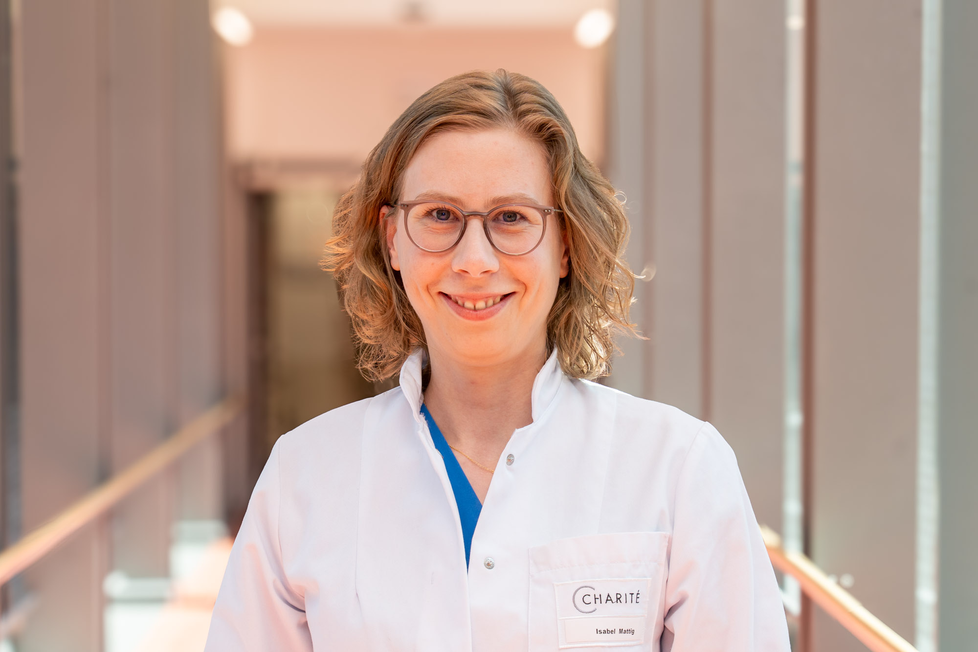 Eine lächelnde Ärztin mit Brille trägt einen weißen Kittel. Sie steht in einem modernen Krankenhausflur mit rotem Licht und grauen Wänden. Ihr Namensschild ist sichtbar. Der Fokus liegt auf ihrer freundlichen Ausdrucksweise und professionellen Erscheinung.