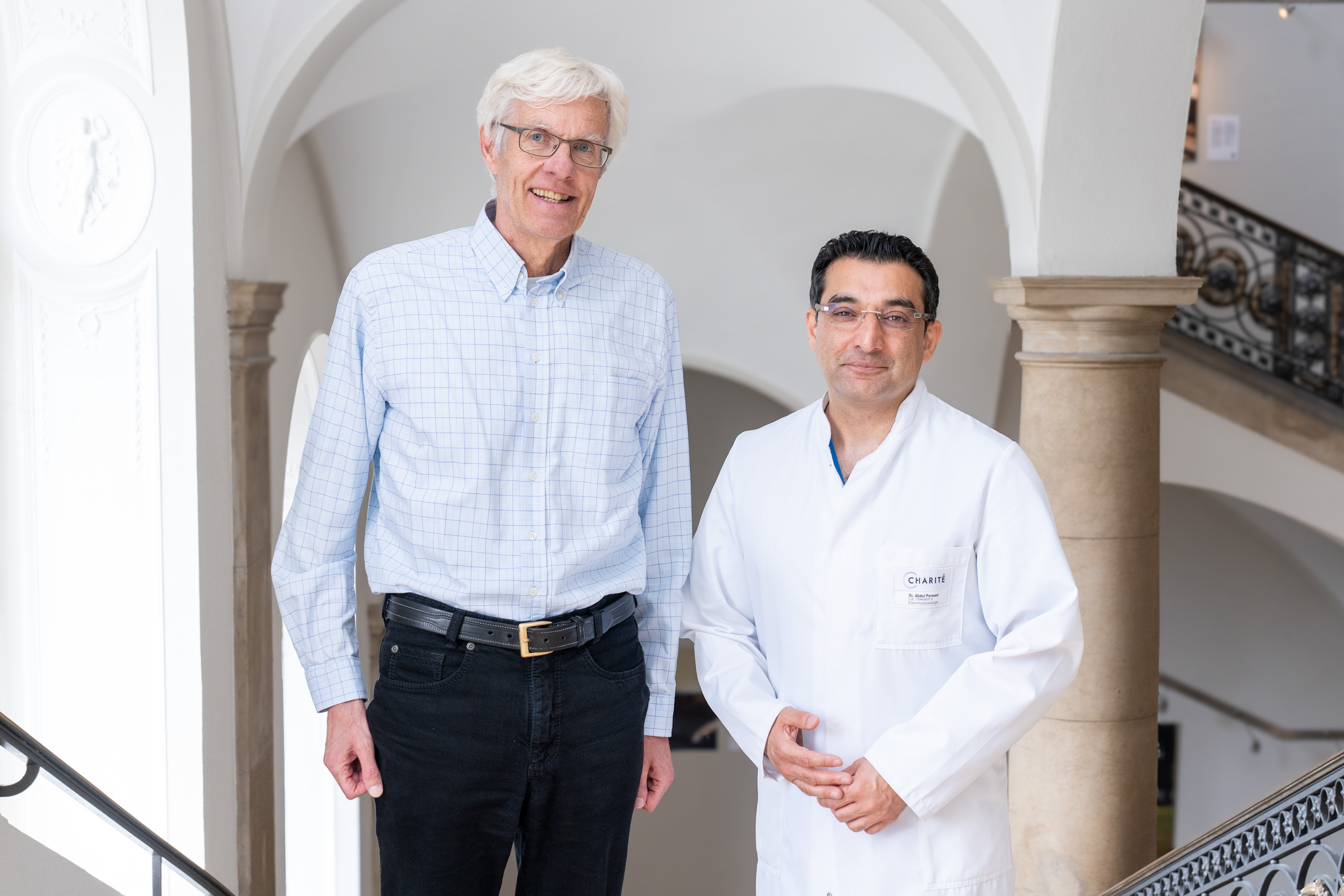 Zwei Männer stehen auf einer Treppe. Der Mann links trägt ein karierter Hemd und Jeans, der Mann rechts ist im weißen Arztkittel. Beide lächeln in die Kamera und stehen vor einem architektonisch ansprechenden Hintergrund.