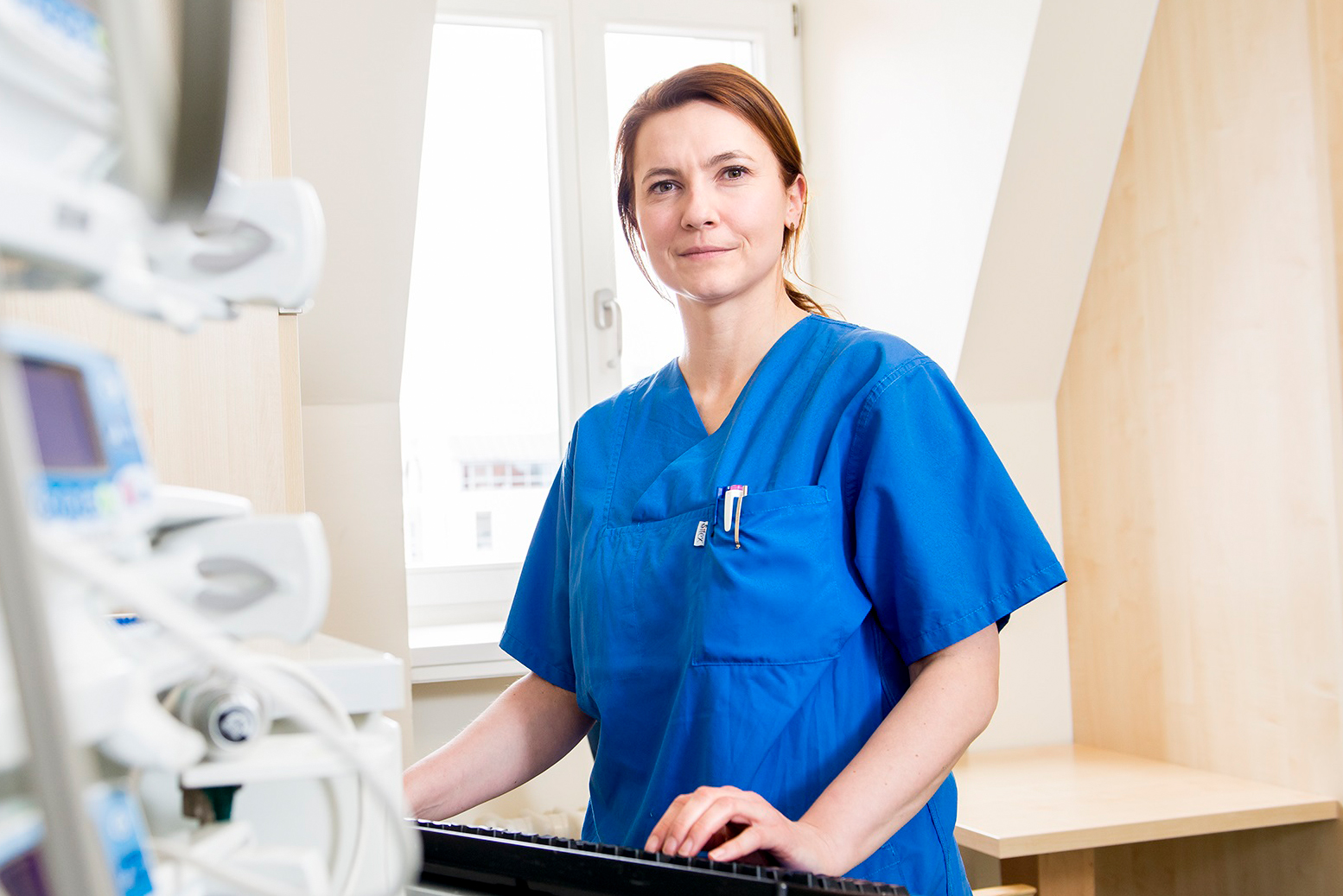Eine Krankenschwester in blauer Arbeitskleidung steht an einem medizinischen Gerät und blickt freundlich in die Kamera. Im Hintergrund sind helle Wände und Fenster sichtbar, die eine ruhige, professionelle Umgebung schaffen.