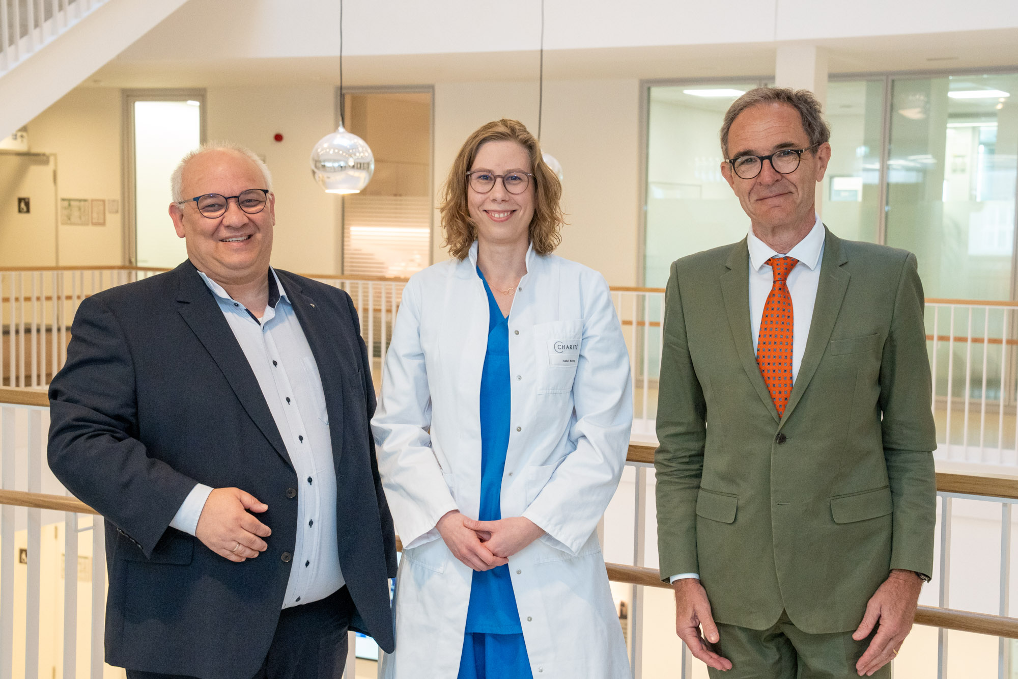 Drei Personen stehen in einem modernen Innenraum. Eine Frau in einem weißen Kittel vermittelt einen medizinischen Kontext, während die anderen beiden Personen in formeller Kleidung gekleidet sind. Alle lächeln und blicken in die Kamera.