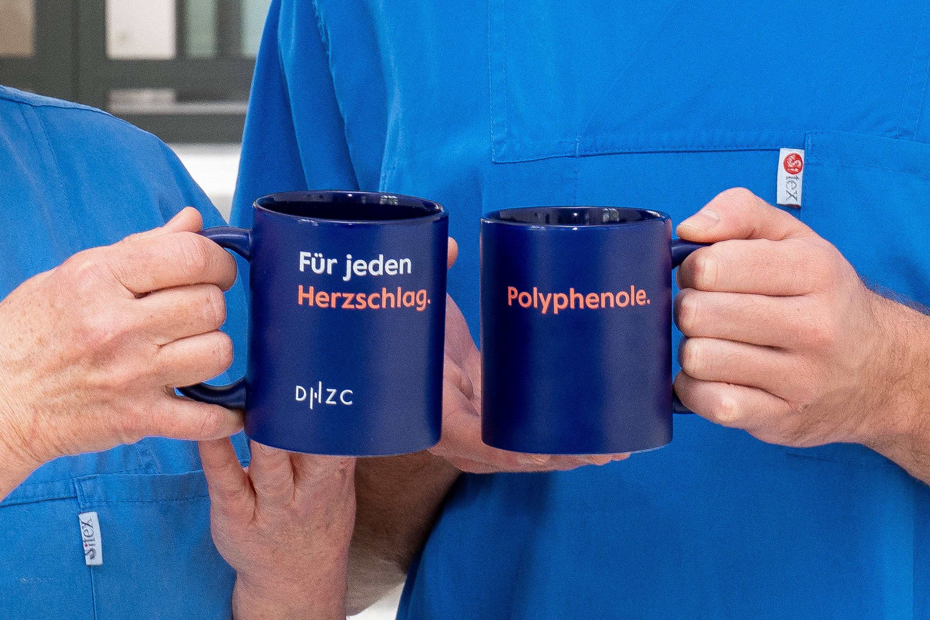 Zwei Personen in blauen Kitteln halten zwei Tassen. Eine Tasse trägt den Text "Für jeden Herzschlag," die andere "Polyphenole." Die Tassen sind dunkelblau und scheinen zu einem gesundheitlichen oder ernährungswissenschaftlichen Thema zu gehören.