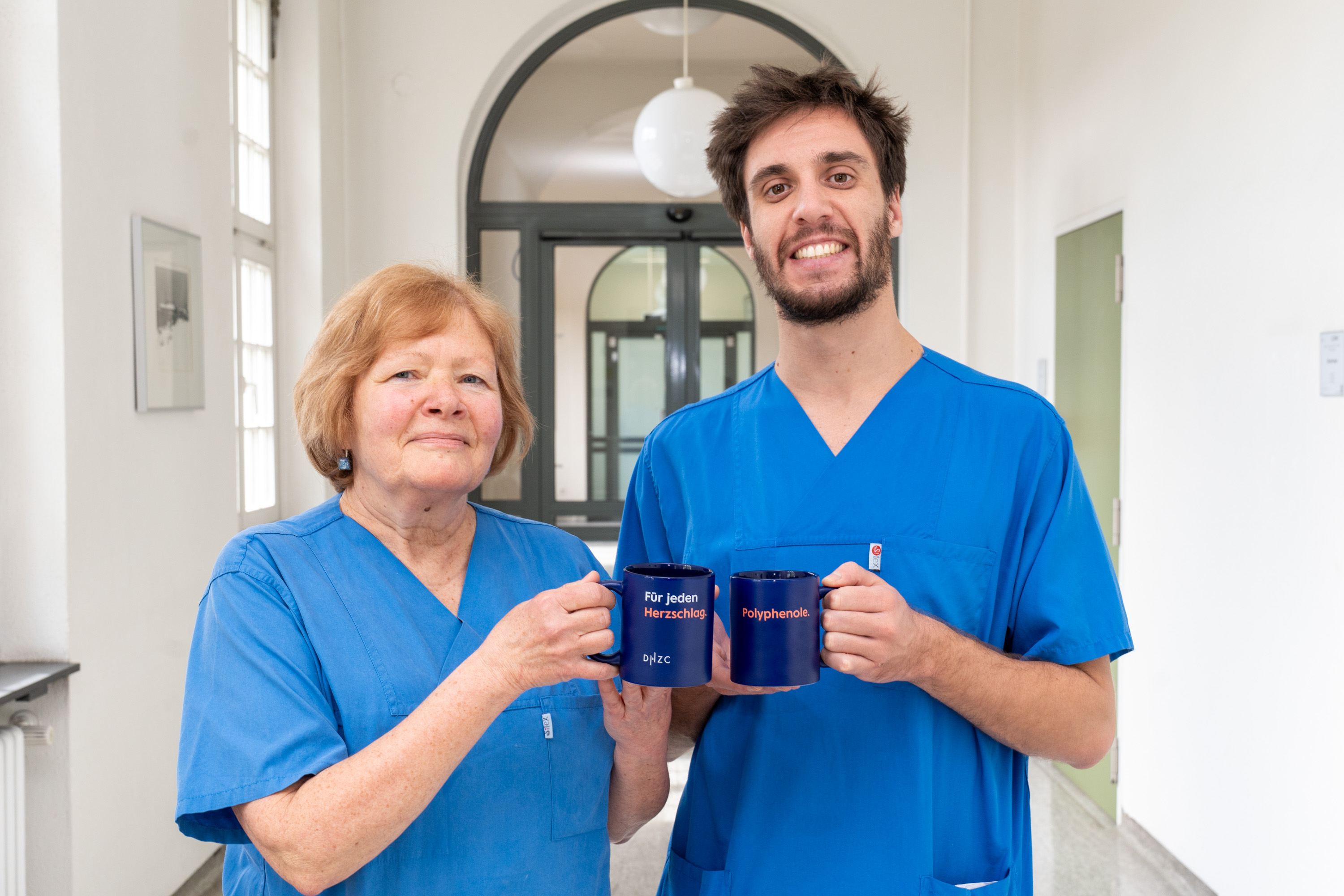 Zwei Personen in blauen Arbeitskleidern stehen in einem Flur und halten jeweils eine Tasse in der Hand. Die Tassen haben aufgedruckte Texte, die Gemeinschaft und Unterstützung symbolisieren. Die beiden Personen lächeln und strahlen eine positive Atmosphäre aus.