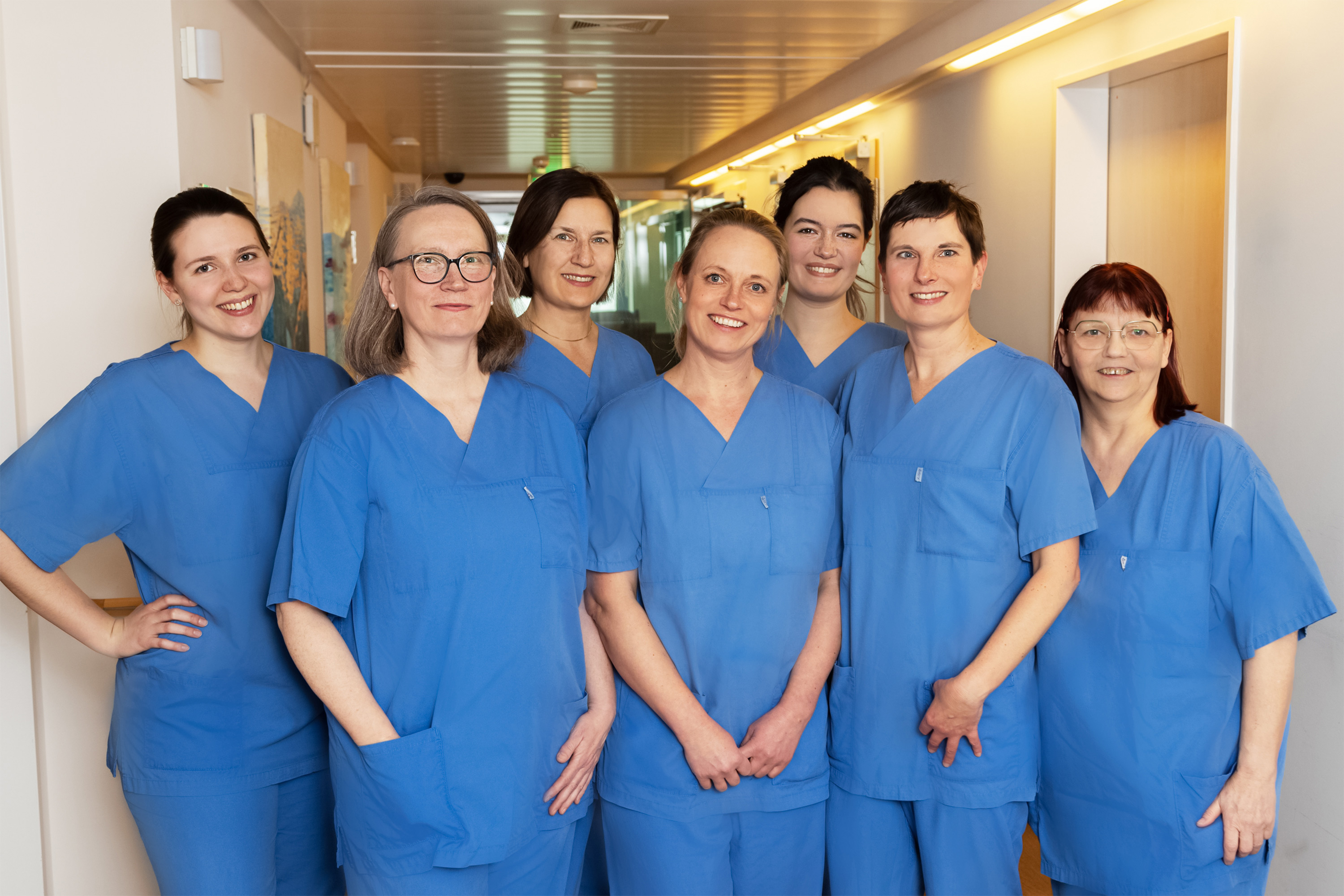 Gruppe von sieben Frauen in blauen Arbeitskleidung, die freundlich in einem Flur stehen. Sie lächeln in die Kamera und strahlen Teamgeist aus. Der Hintergrund zeigt eine helle Umgebung, die auf ein medizinisches oder pflegerisches Umfeld hinweist.
