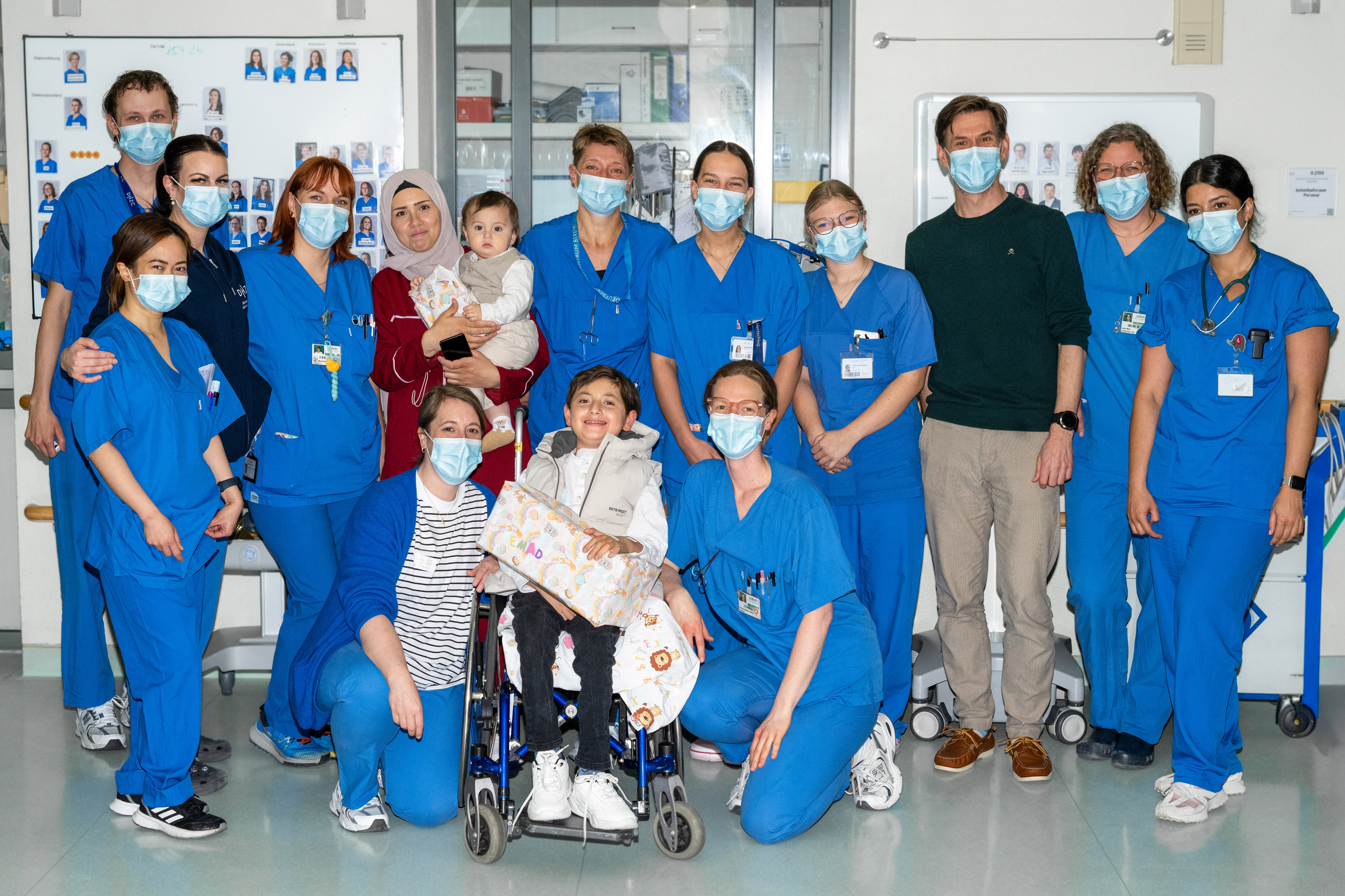 Eine Gruppe von 16 Personen in blauen Krankenhausuniformen steht in einem Raum. Einige halten Kinder, während andere lächeln und den Betrachter anblicken. Im Vordergrund sitzt ein Kind im Rollstuhl, umgeben von Betreuern. Die Atmosphäre ist freundlich und unterstützend.