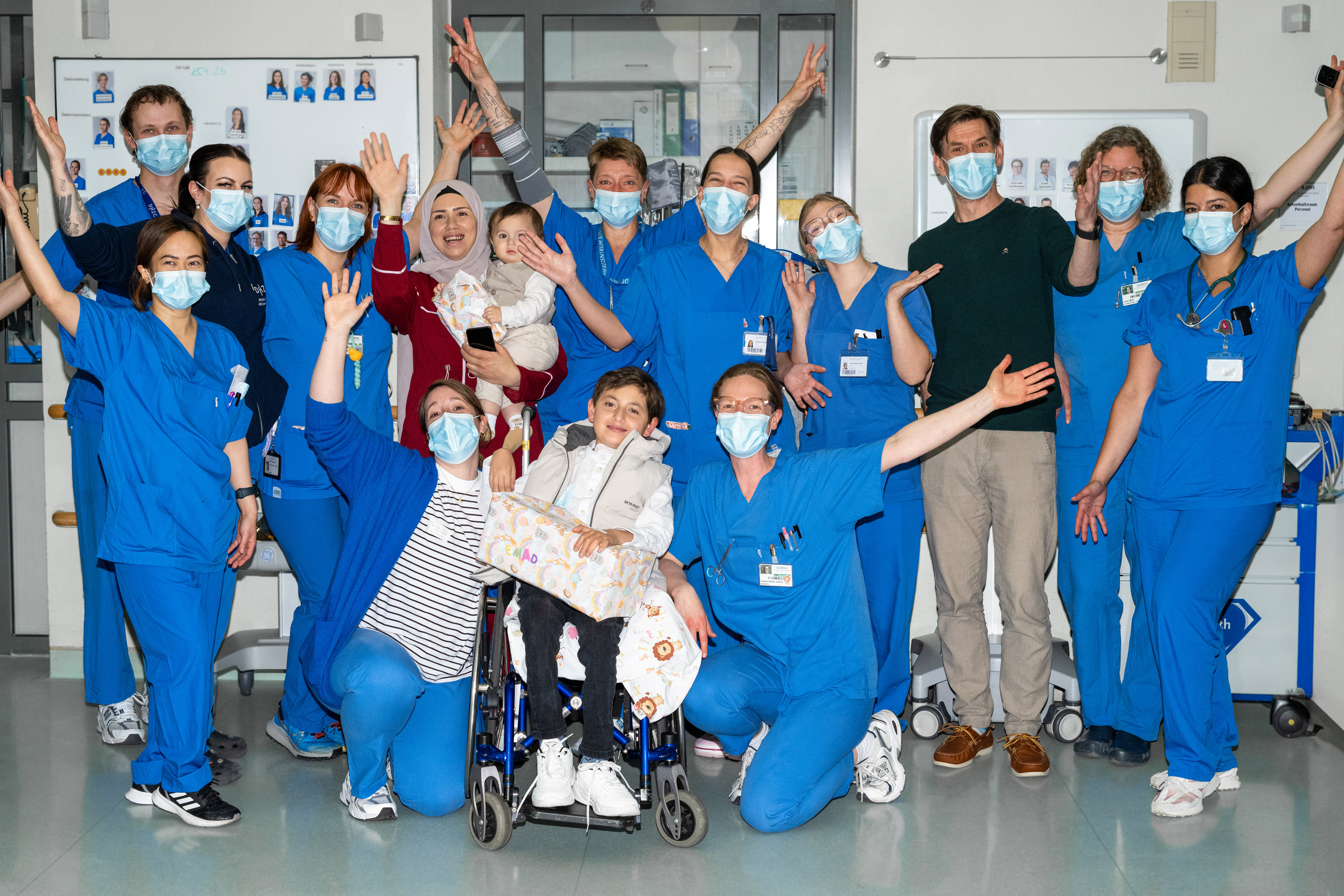 Eine Gruppe von mehr als 15 Krankenhausmitarbeitern in blauen Scrubs und mit Mundschutz posiert fröhlich. In der Mitte sitzt ein Kind im Rollstuhl, umgeben von Angehörigen und Pflegekräften, die ihre Hände in die Luft heben und lächeln. Die Szene strahlt Freude und Teamgeist aus.