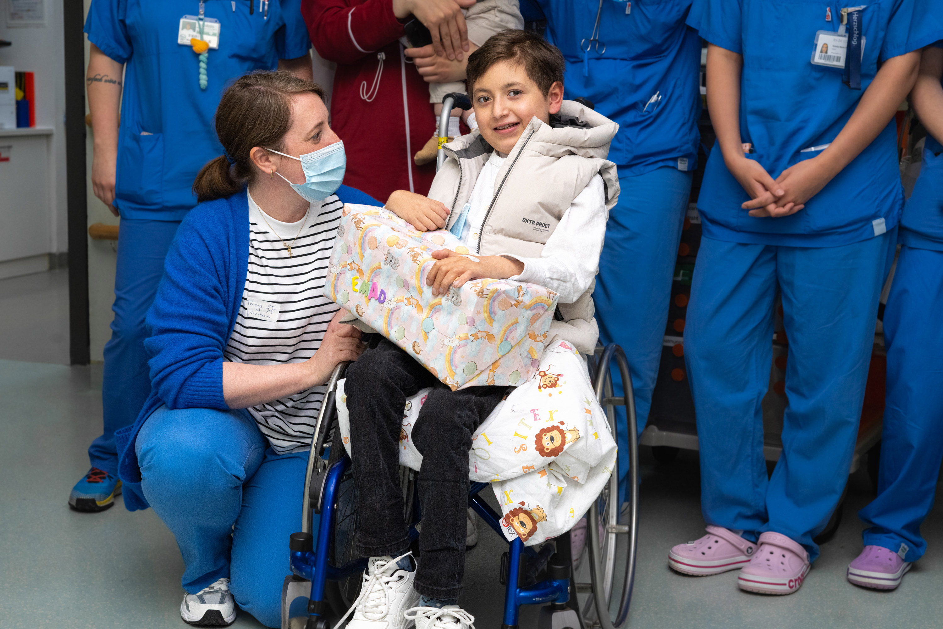 Ein lächelnder Junge in einem Rollstuhl hält ein Geschenk in den Händen. Neben ihm kniet eine Frau in blauer Kleidung und Maske. Im Hintergrund stehen mehrere Personen in blauer Arbeitskleidung. Die Szene strahlt Freude und Unterstützung aus.