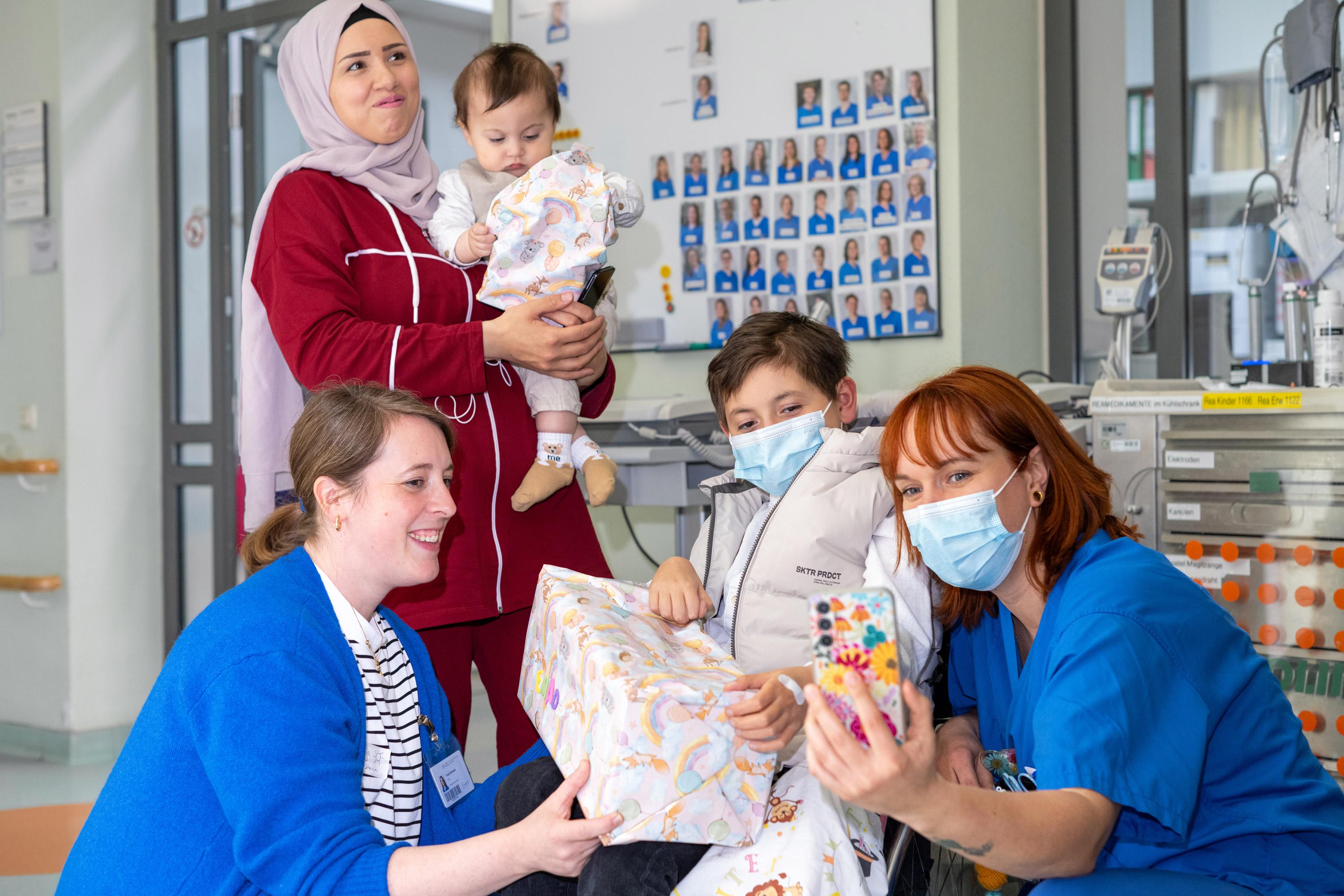 Fünf Personen in einem Krankenhaus: Eine Frau mit Kopfbedeckung hält ein Kleinkind, während ein Junge mit einer Maske ein Geschenk erhält. Zwei weitere Frauen, die ebenfalls Masken tragen, unterstützen und beobachten die Szene. Die Atmosphäre ist freundlich und fürsorglich.